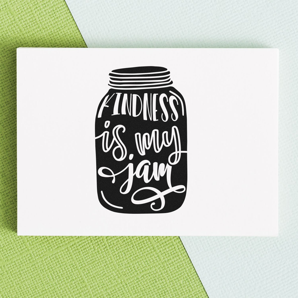 Black jar-shaped stamp seal with Kindness is my jam in white script on a white card, on green and white textured background.