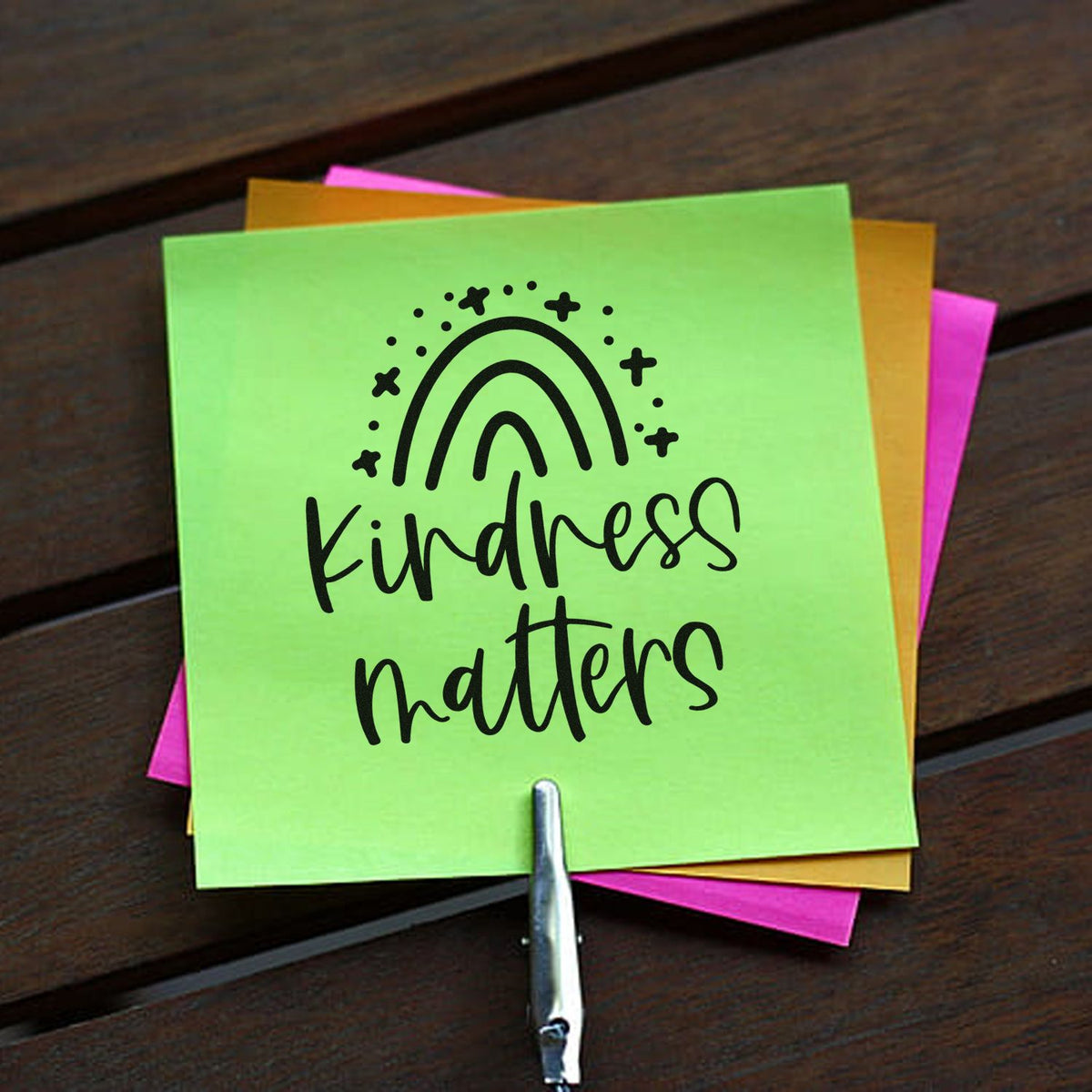 Lime green stamp seal with black rainbow and Kindness matters, layered over pink and yellow notes on wood.