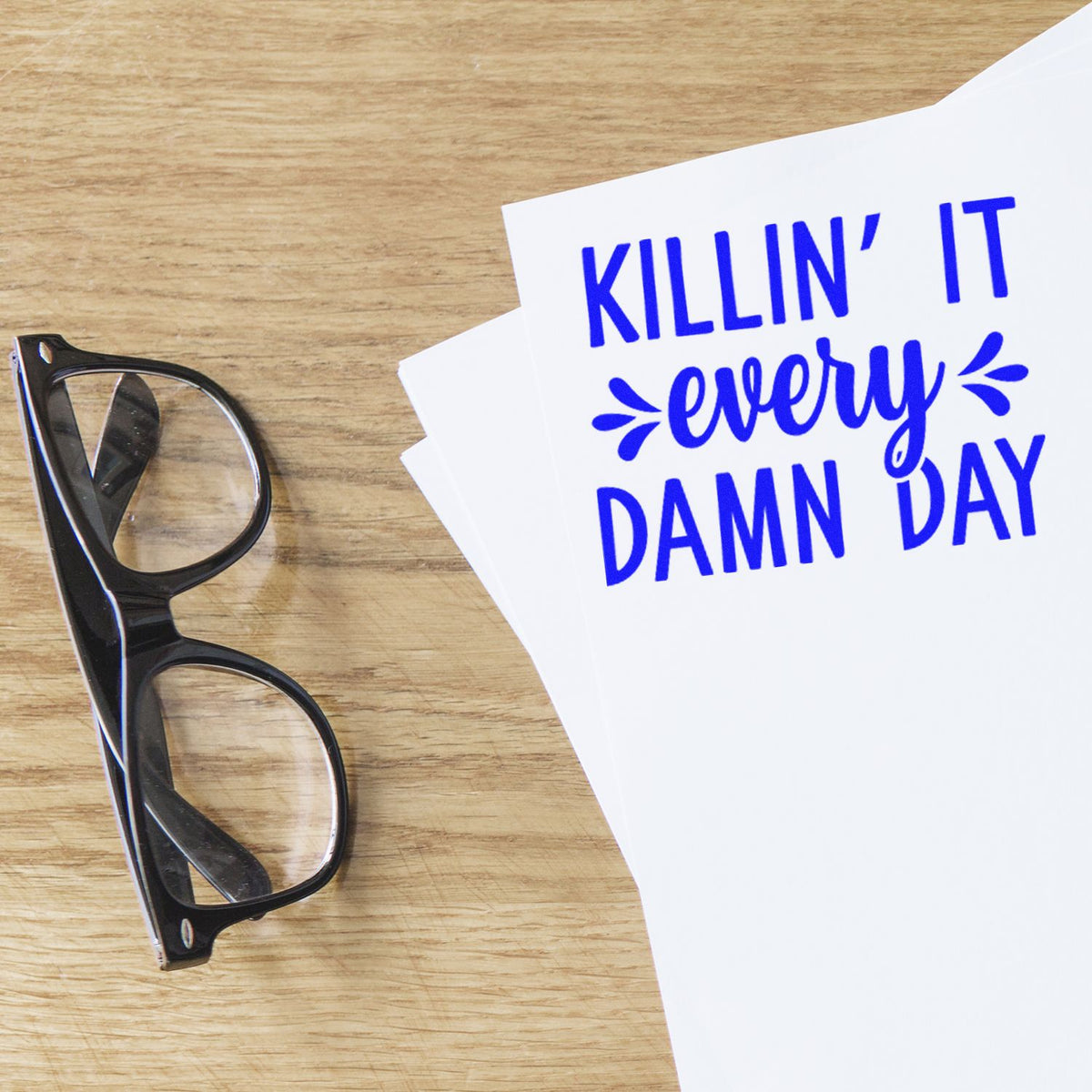 Blue stamp/seal design reading KILLIN IT every DAMN DAY on white paper beside black glasses on a wood surface.