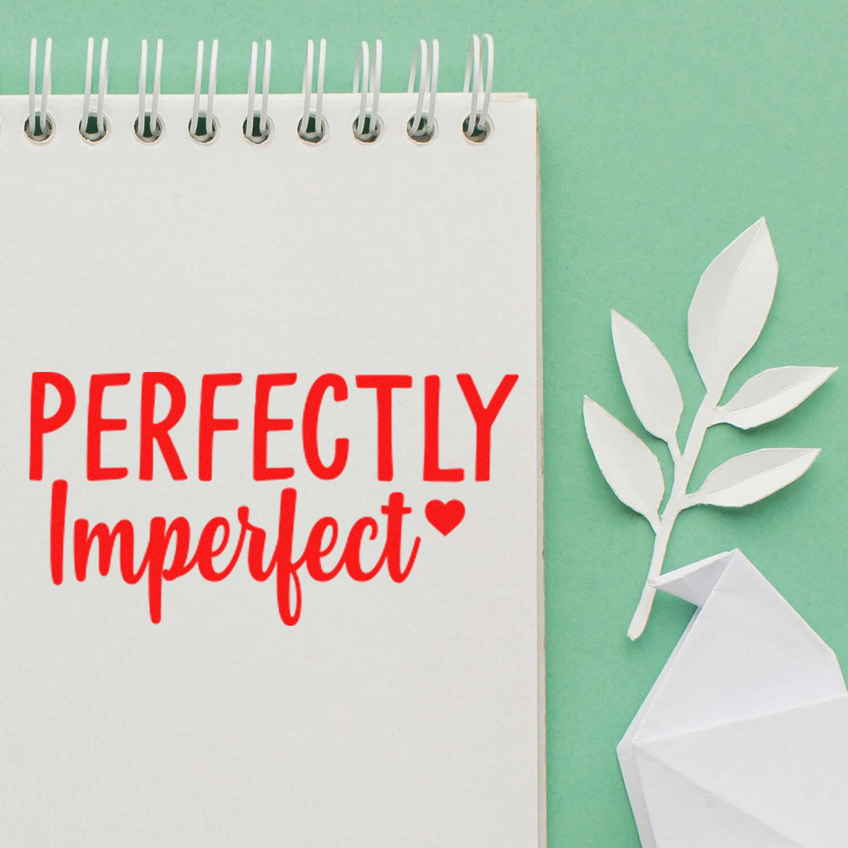 Red Perfectly Imperfect stamp on a white spiral notebook, with white leaf seal and folded paper accents on a mint green background.