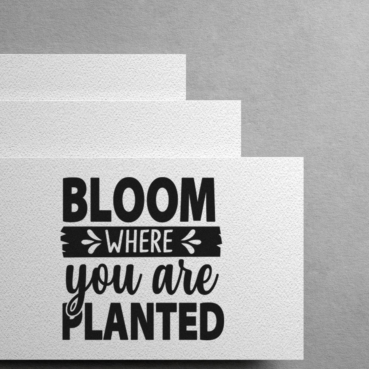 Black stamp-style seal on white textured paper reading Bloom where you are planted with decorative leaf accents.