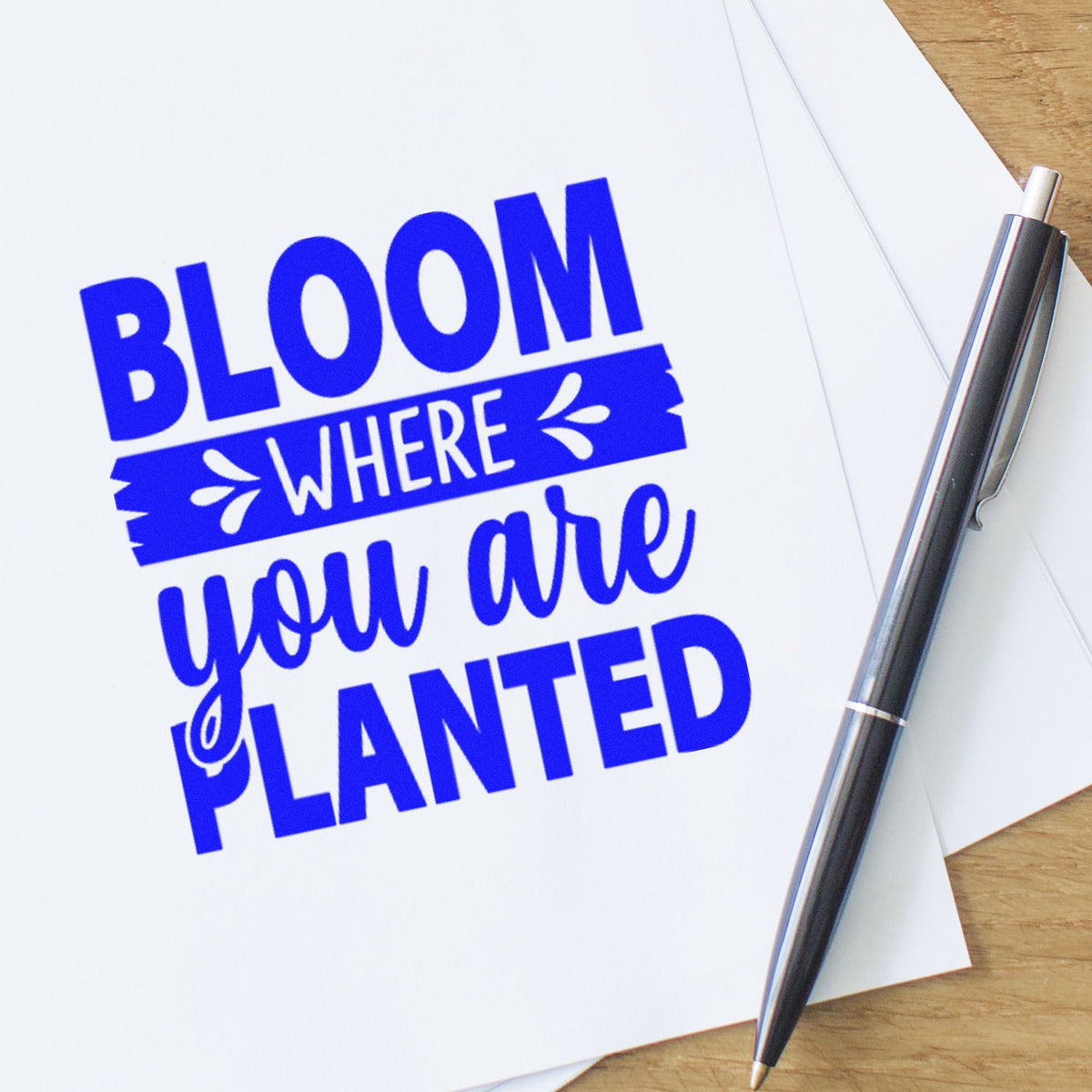 Blue self-inking stamp reading Bloom where you are planted beside a black metal seal pen on white paper.