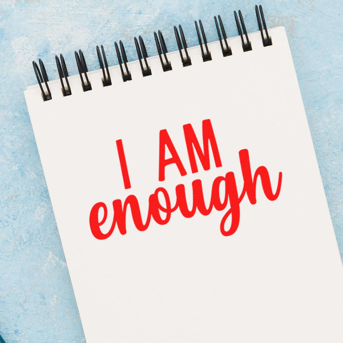 White spiral notepad with red I AM enough stamp-style seal on a blue textured background.