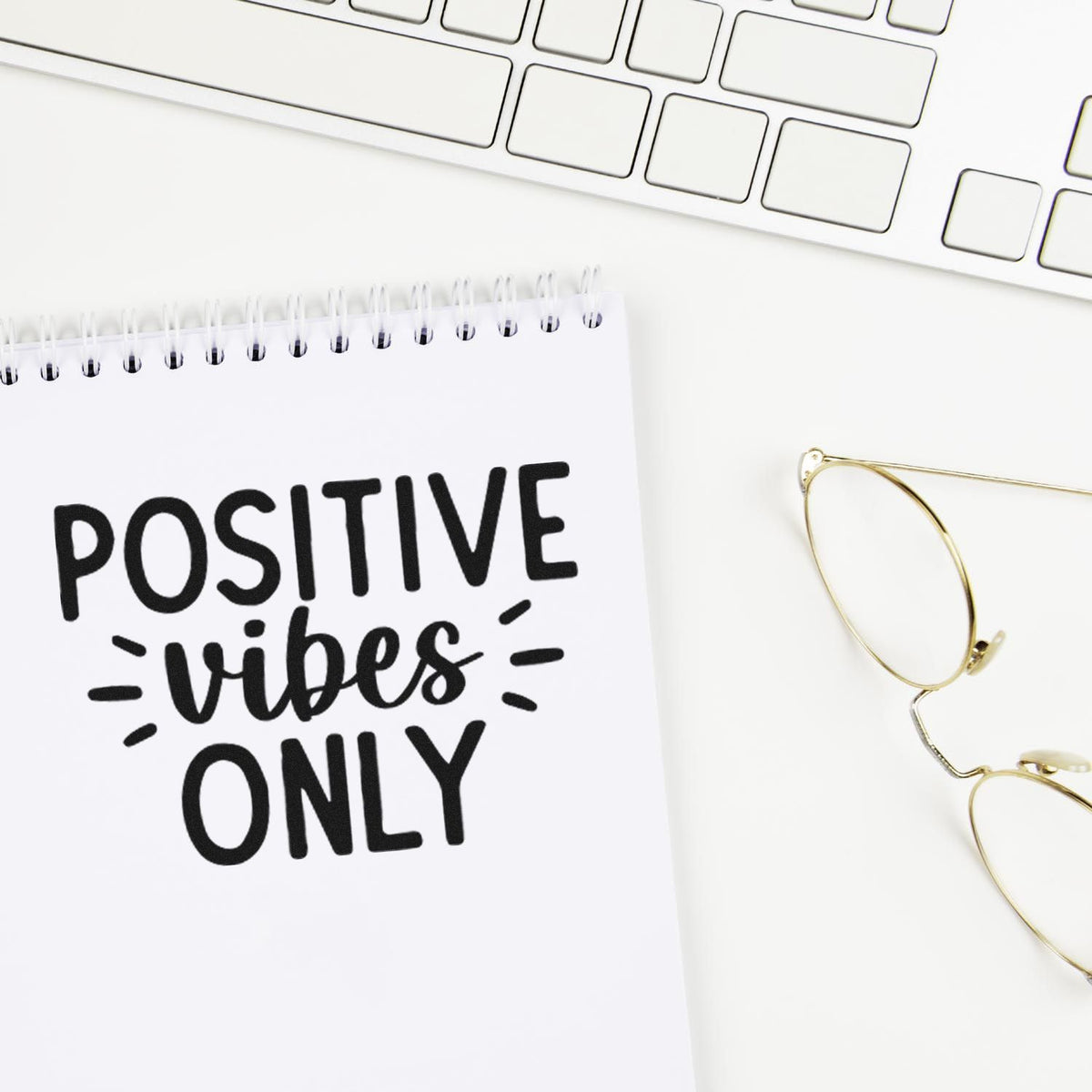 Black Positive Vibes Only stamp on a white spiral notebook, with a keyboard and gold glasses.