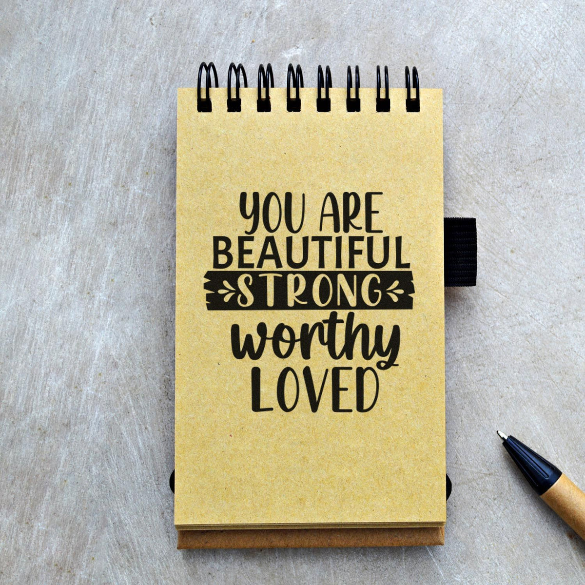Yellow spiral stamp-and-seal notebook with black You are beautiful, strong, worthy, loved text on a gray tabletop.