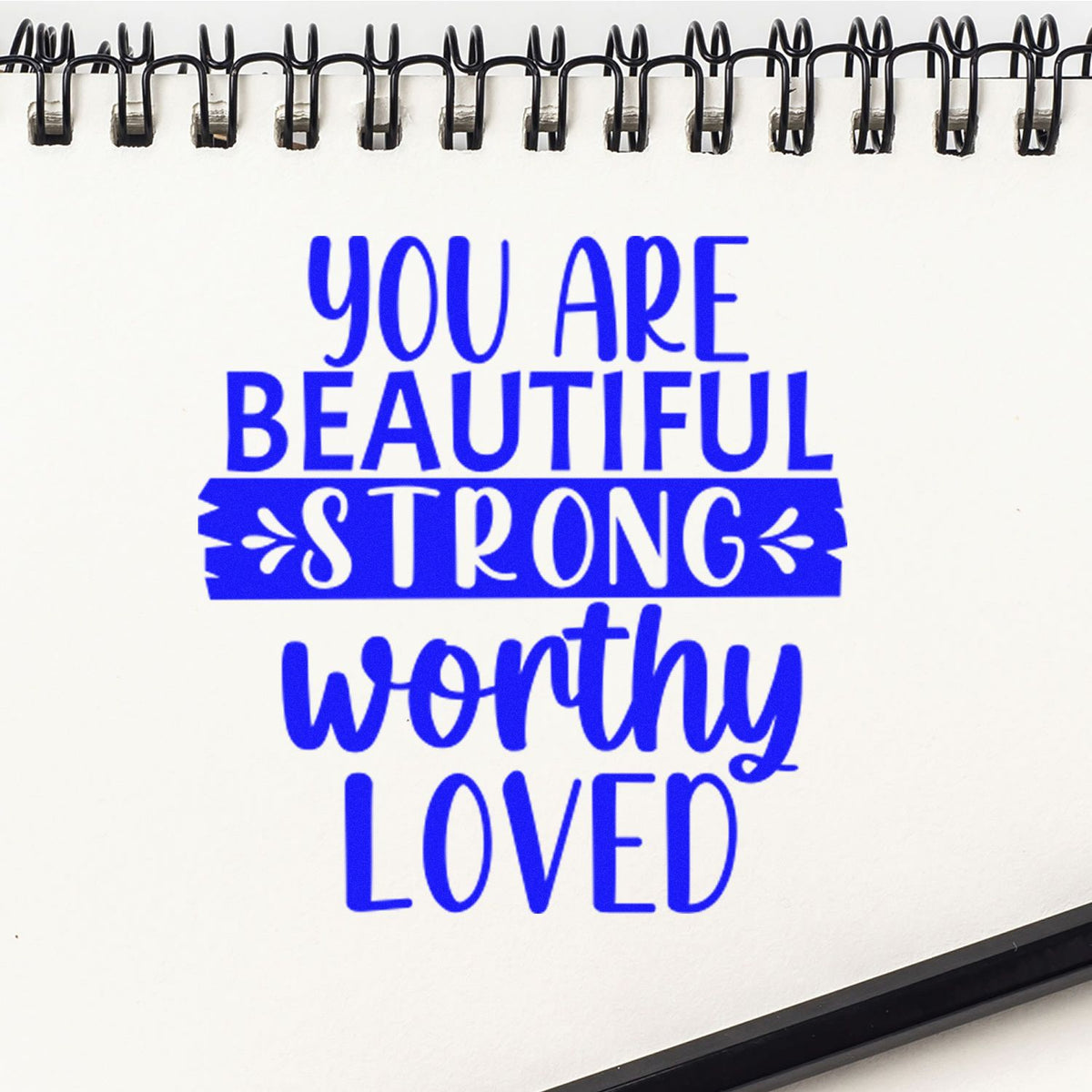 Blue motivational rubber stamp and seal on white spiral notebook: You are beautiful, strong, worthy, loved.