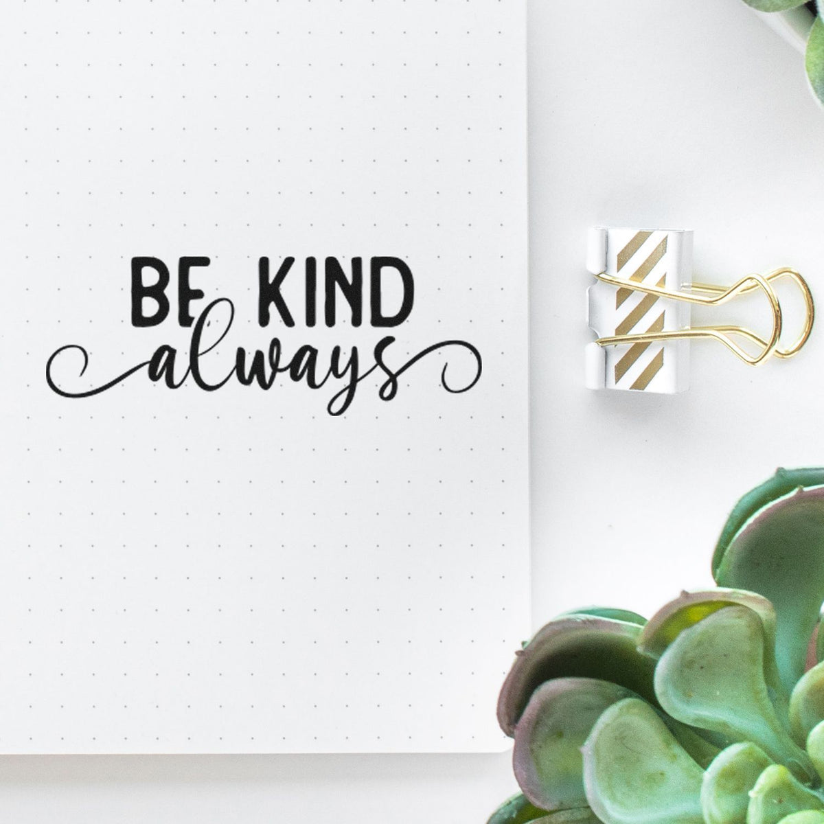 White dotted notebook page with black BE KIND always stamp, gold-striped binder clip, and green succulent accents.