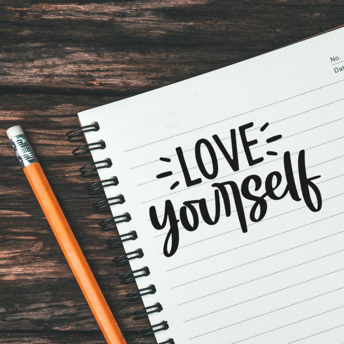 Black LOVE yourself stamp on a white spiral notebook, with an orange pencil on a dark wood background.