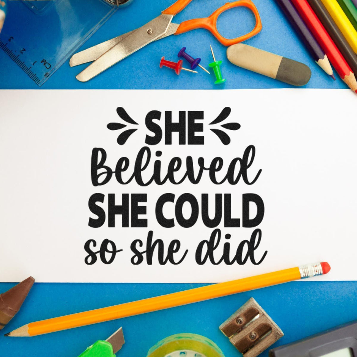 Black stamp quote on white paper, surrounded by blue school supplies and seal tools: She believed she could so she did.