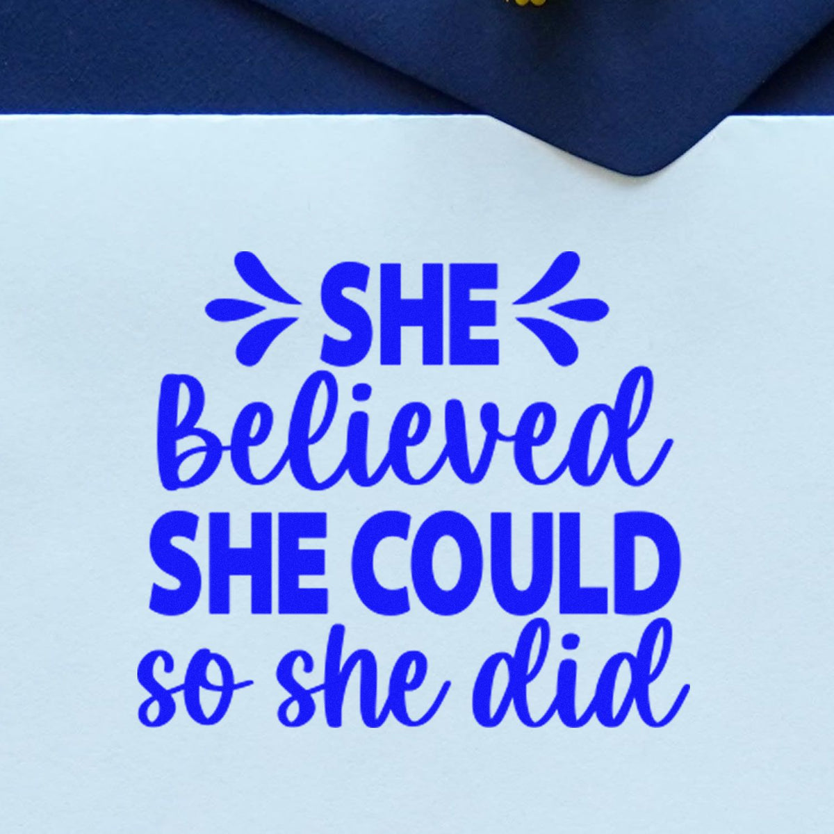 Blue self-inking stamp seal with quote She Believed She Could So She Did in bold blue on white paper.