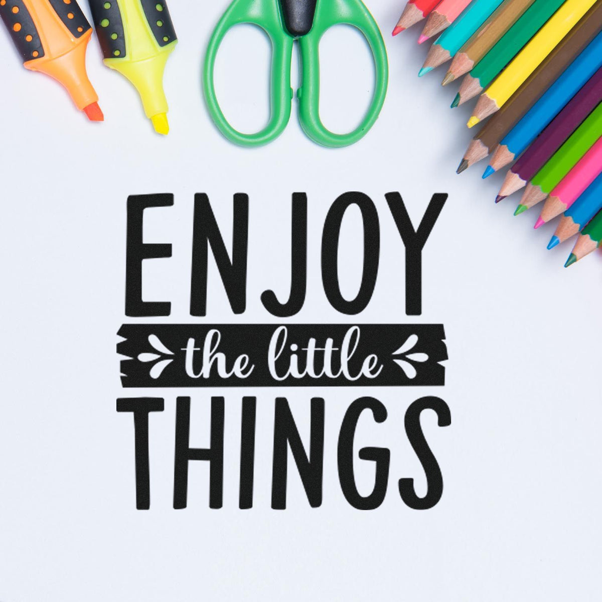 Black stamp and seal design reading ENJOY the little THINGS, framed by colorful pencils, markers, and green scissors on white.