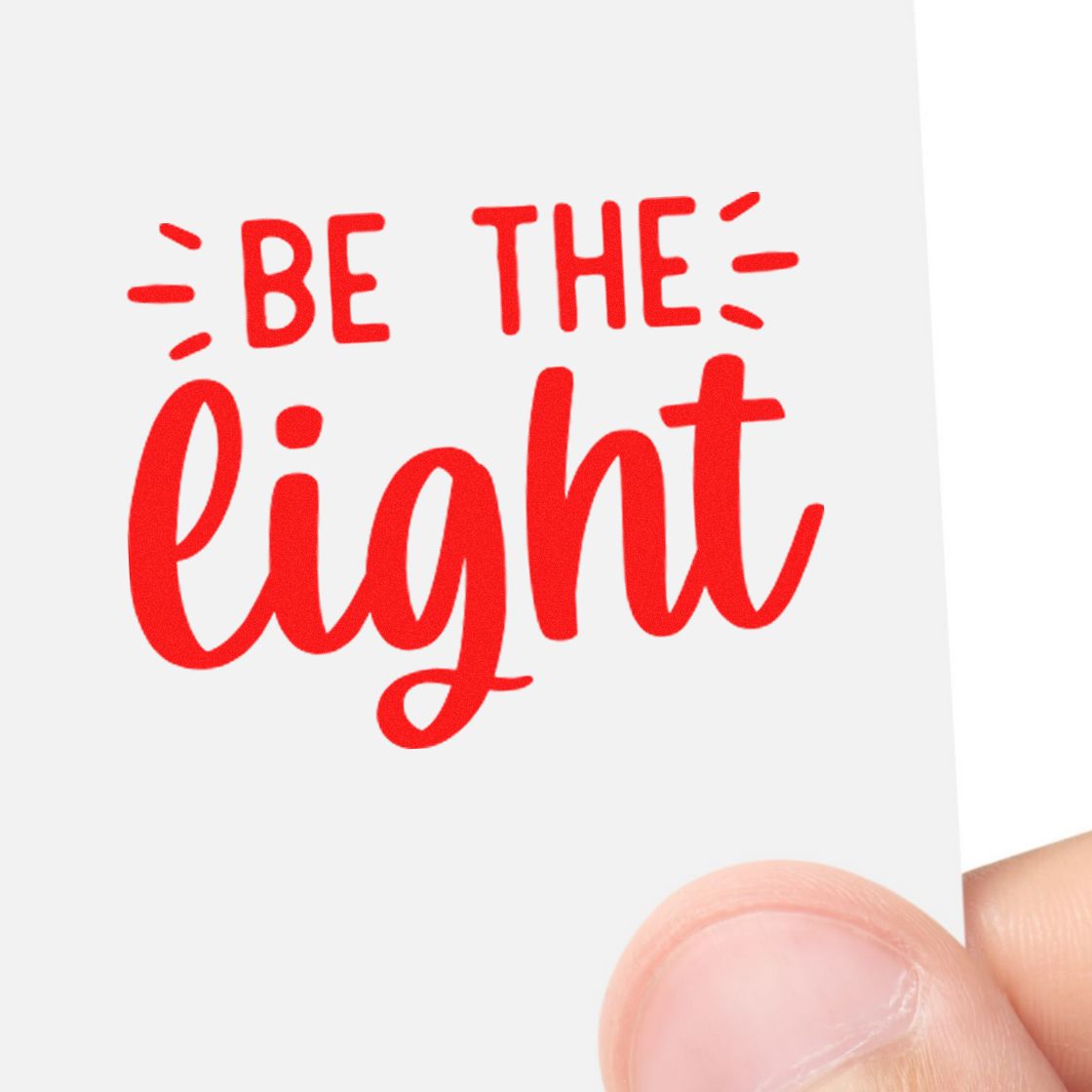 Red rubber stamp reading Be the Light in bright red on a white card, held by hand.