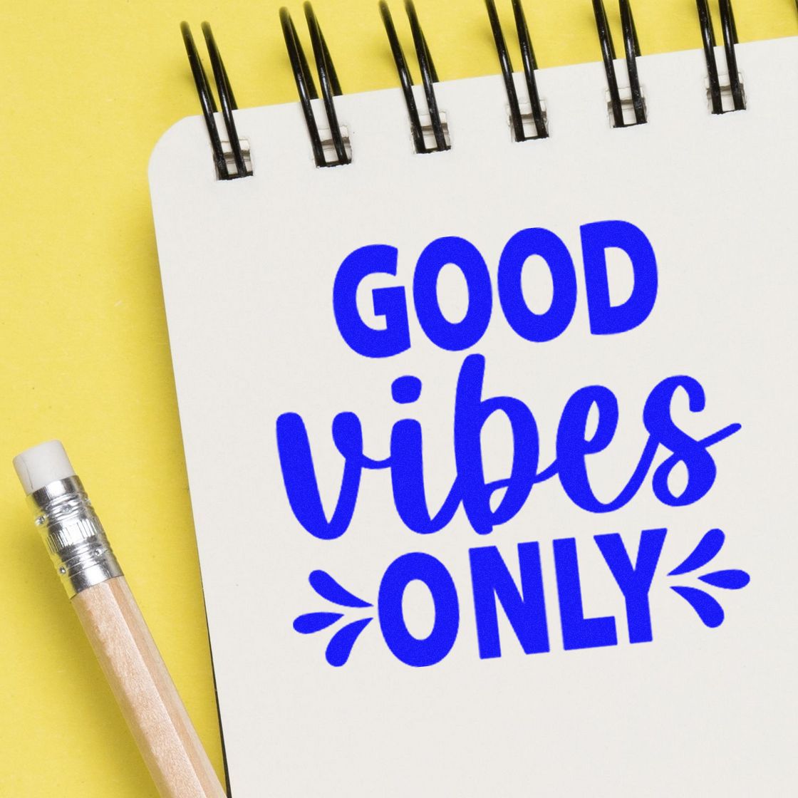 Blue Good Vibes Only stamp design on a white spiral notebook over a yellow background, with a pencil.