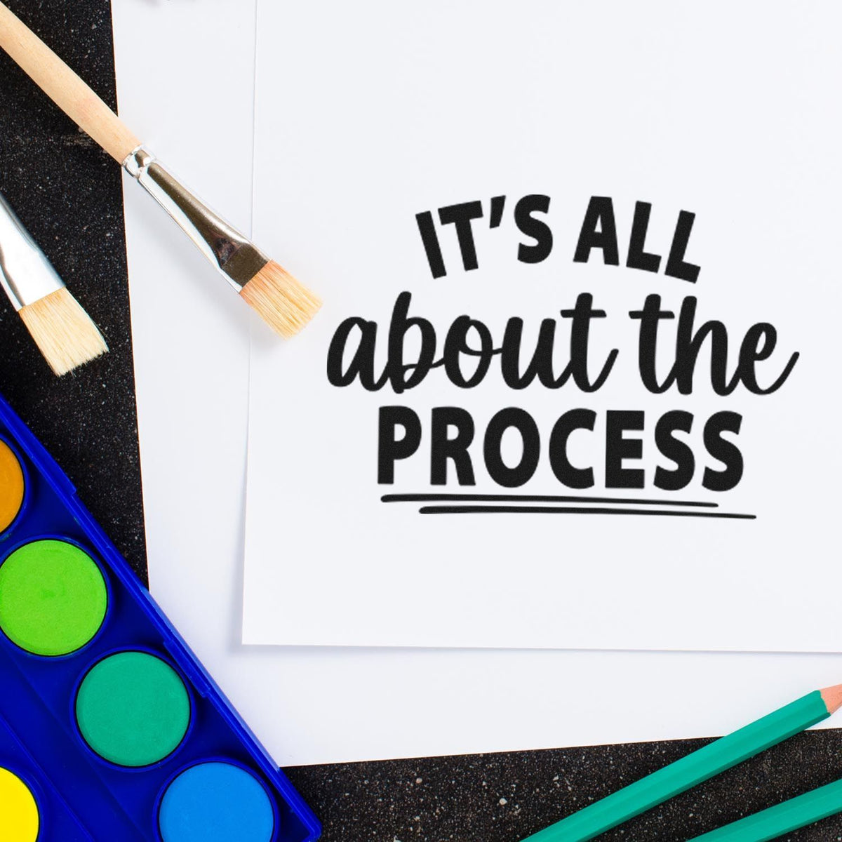 Black stamp reading Its all about the process on white paper, with paintbrushes, teal pencils, and a colorful watercolor palette.