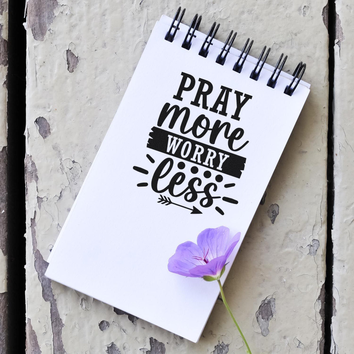 White spiral notebook with black Pray more worry less stamp and seal design, accented by a purple flower.
