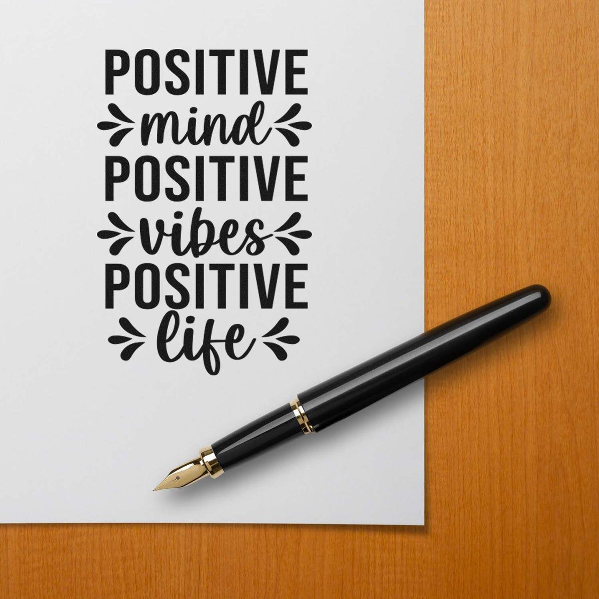 Black decorative stamp reading Positive mind, positive vibes, positive life on white paper, with black fountain pen on wood desk.