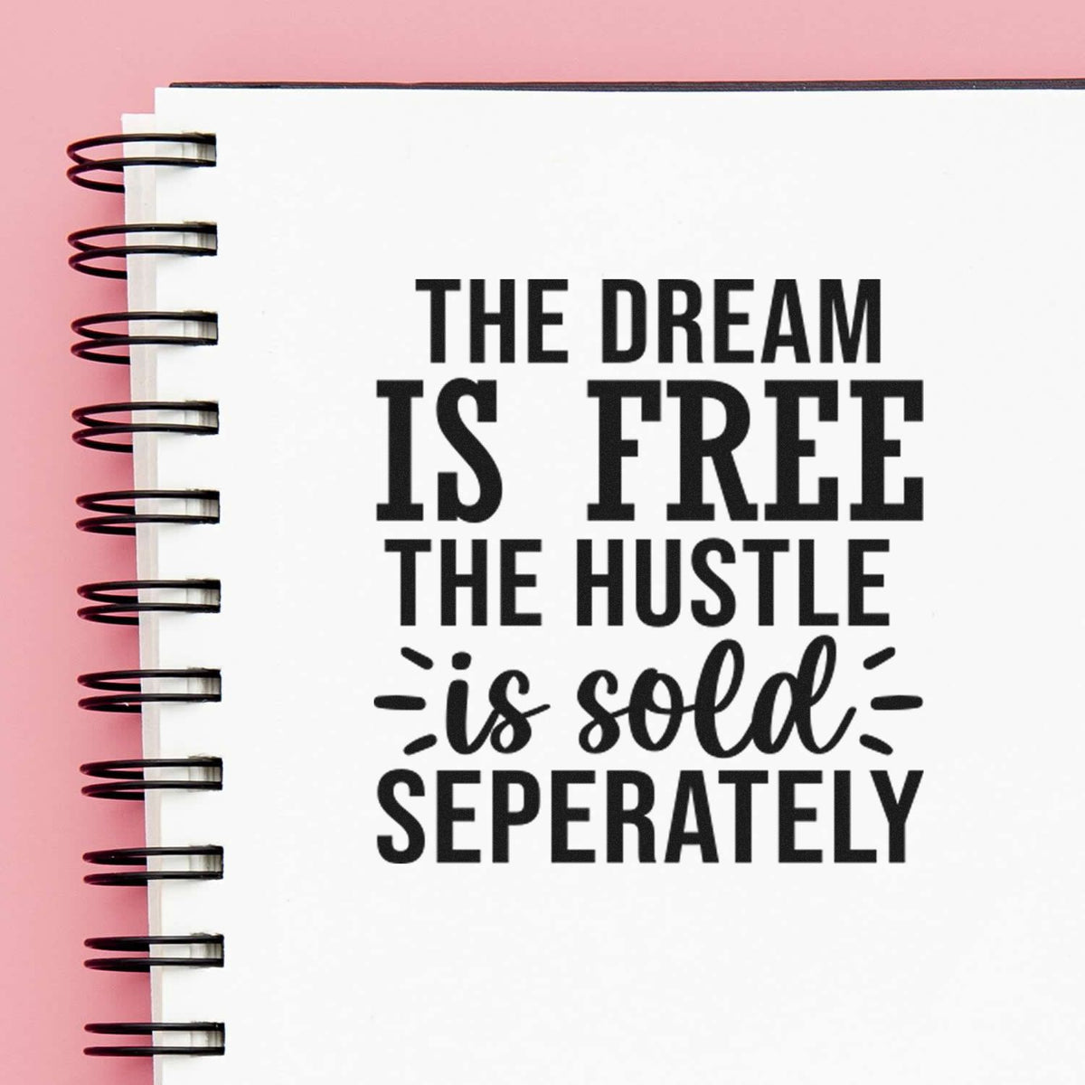 Black stamp-style seal text on white spiral notebook with pink background: The dream is free, the hustle is sold separately.