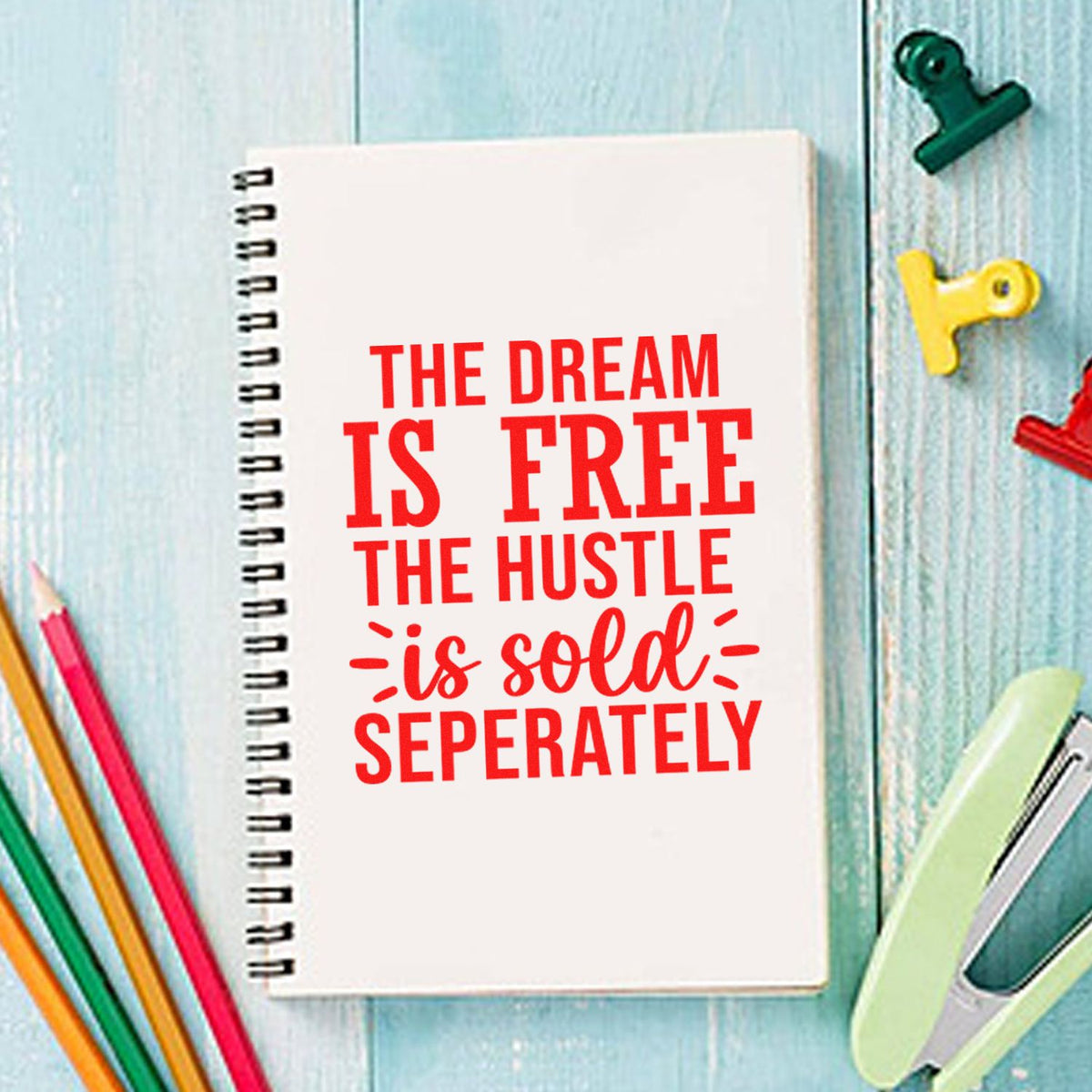 Spiral notebook with red slogan, surrounded by colorful stamp and seal clips on a light blue wood background.