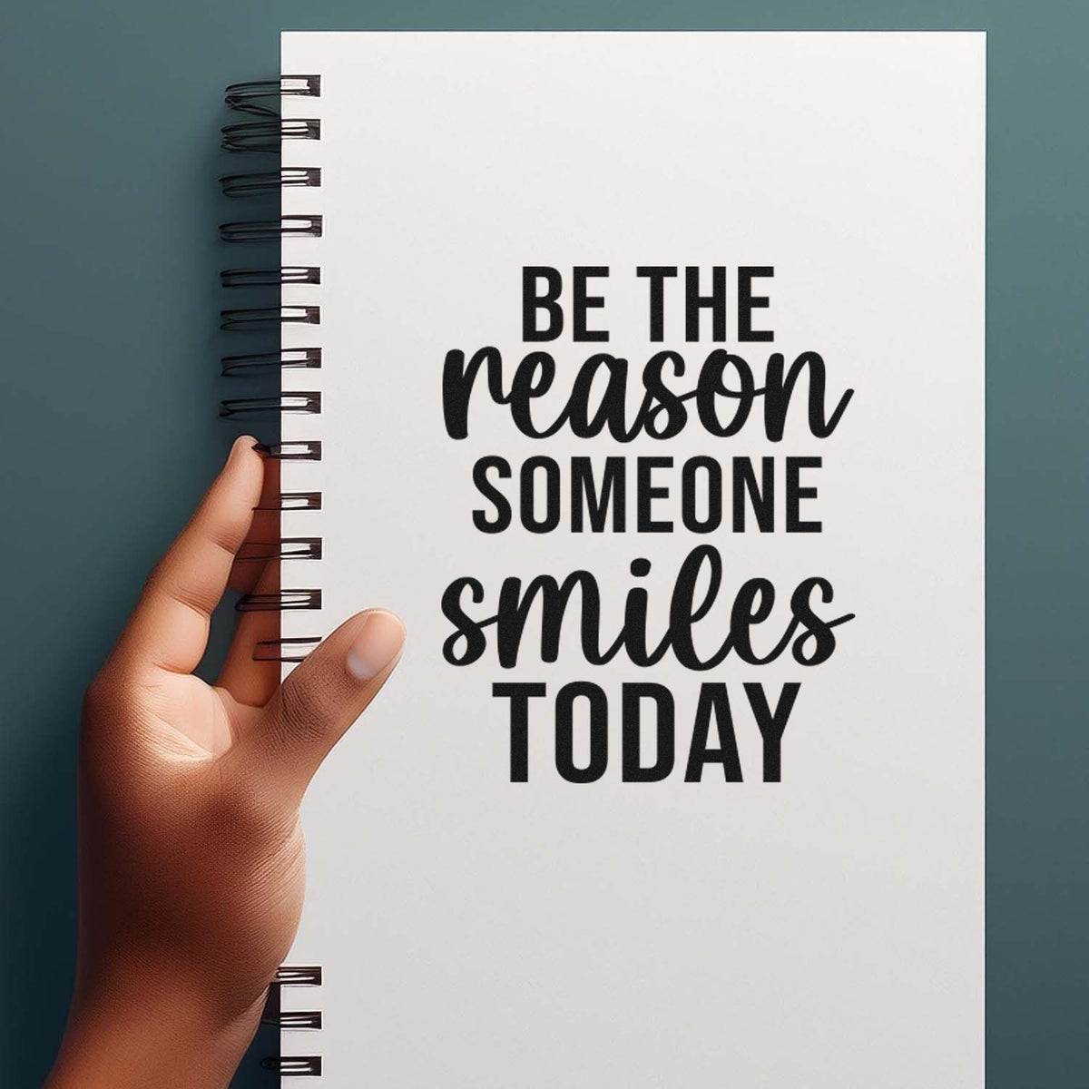 White spiral notebook with black stamp-style quote Be the reason someone smiles today on teal background, held in hand.
