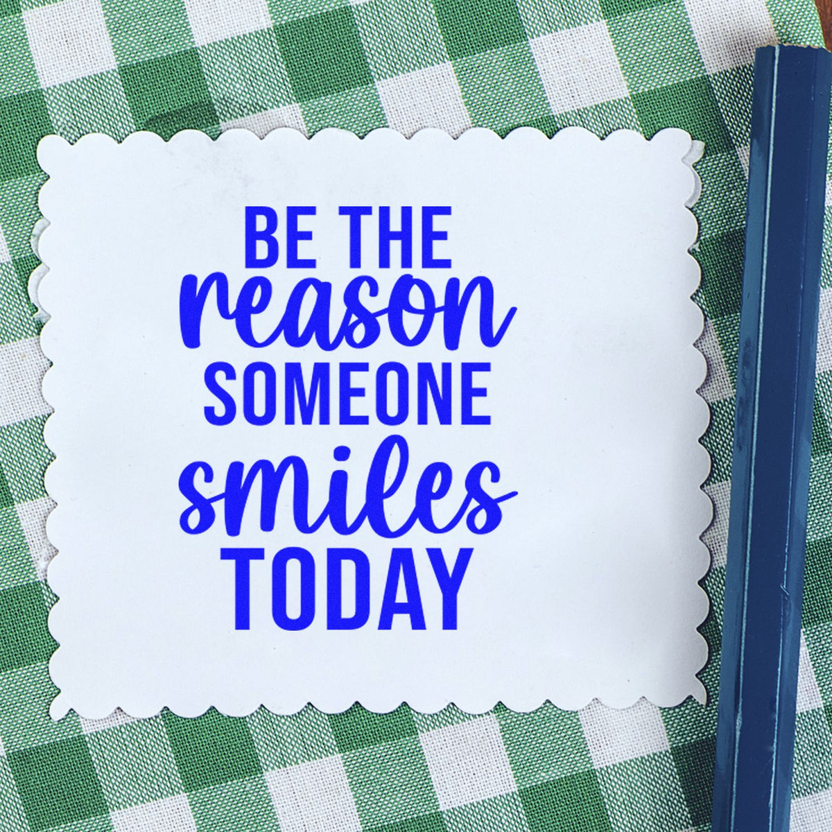 Blue stamp seal with scalloped white label reading Be the reason someone smiles today on green gingham background.