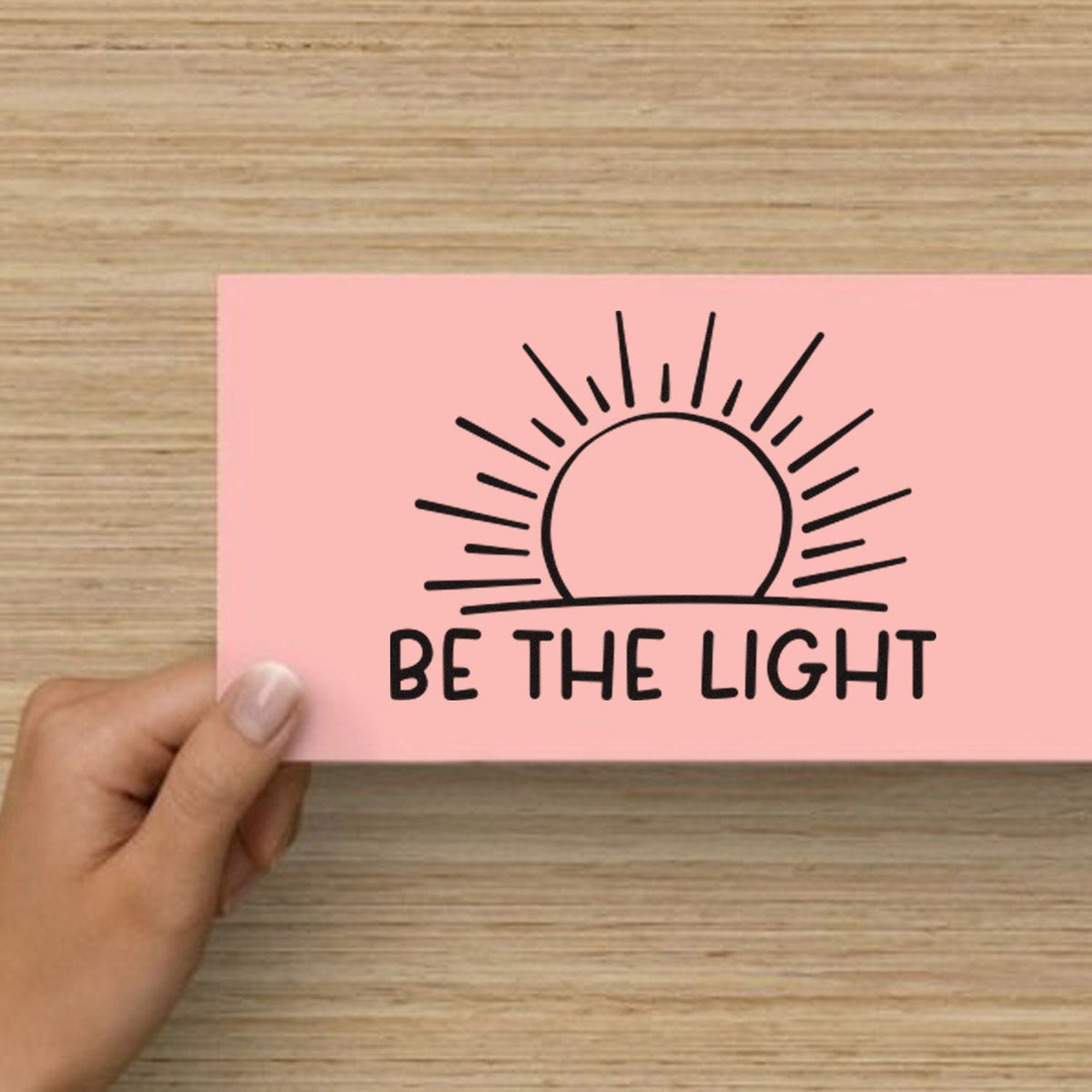 Pink rubber stamp seal card with black sunrise design and BE THE LIGHT text, held on a wood table.