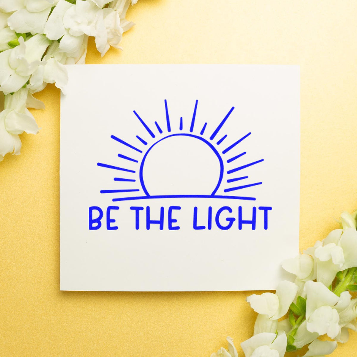 Blue Be The Light stamp and seal with sunburst on white card, on a yellow background with white flowers.