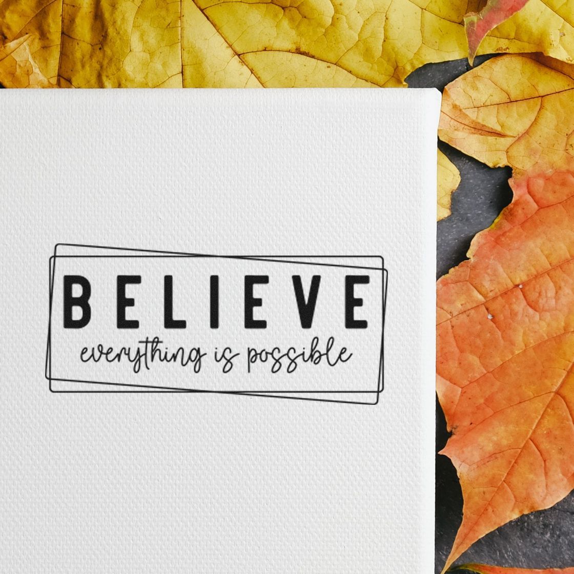 Black self-inking stamp seal reading BELIEVE everything is possible on white paper with yellow and orange autumn leaves.