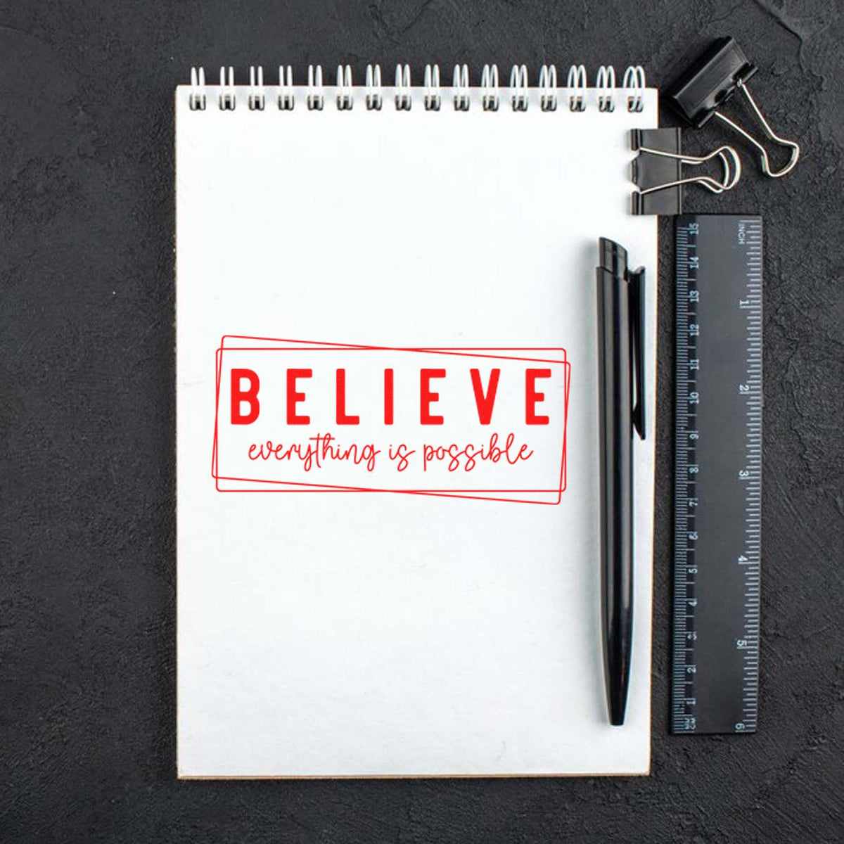 Red motivational stamp and seal reading BELIEVE everything is possible on a white spiral notebook with black pen and ruler.