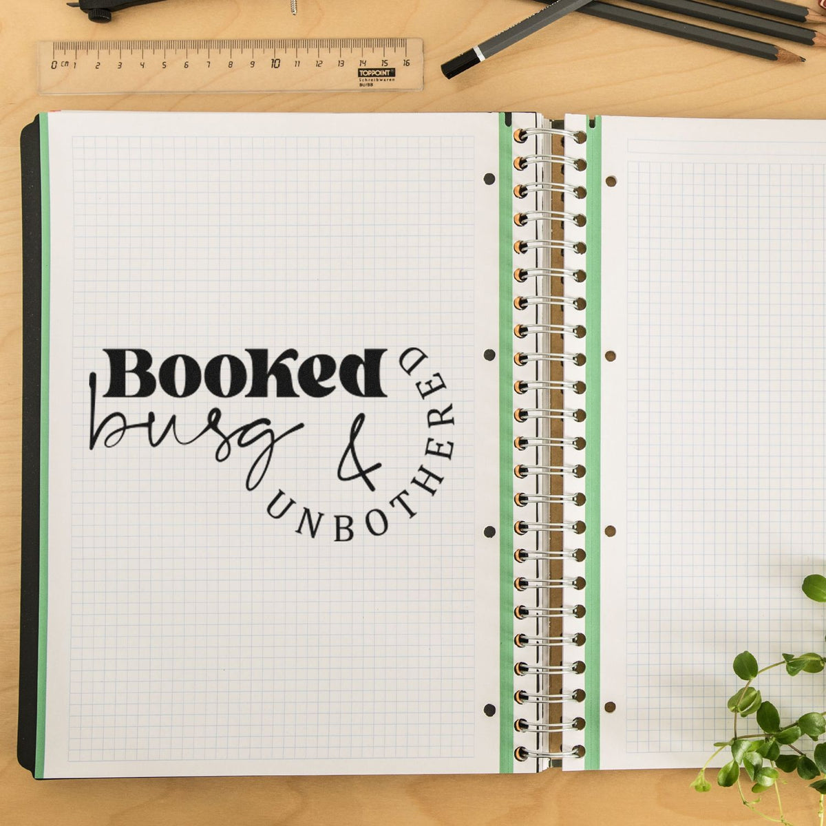 Black stamp and seal branding on an open graph notebook with spiral binding, green edges, ruler, pencils, and wood desk.
