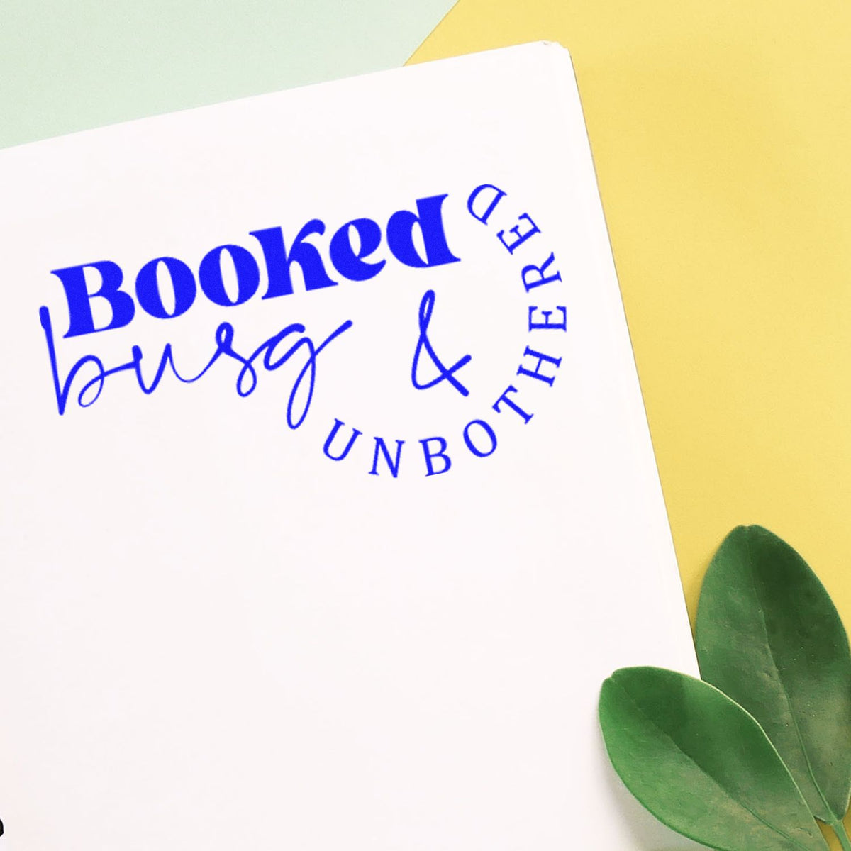 Blue Booked busy & unbothered stamp seal on white paper, with a yellow background and green leaves.