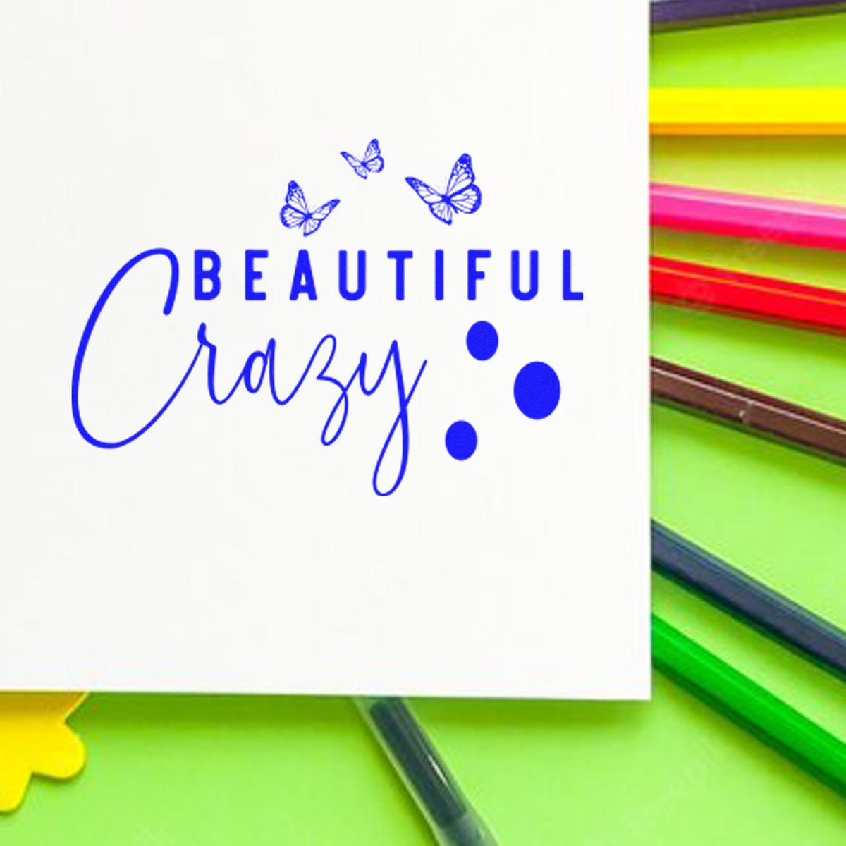 Blue Beautiful Crazy stamp and seal design with butterflies and dots, beside colorful pens on a green background.