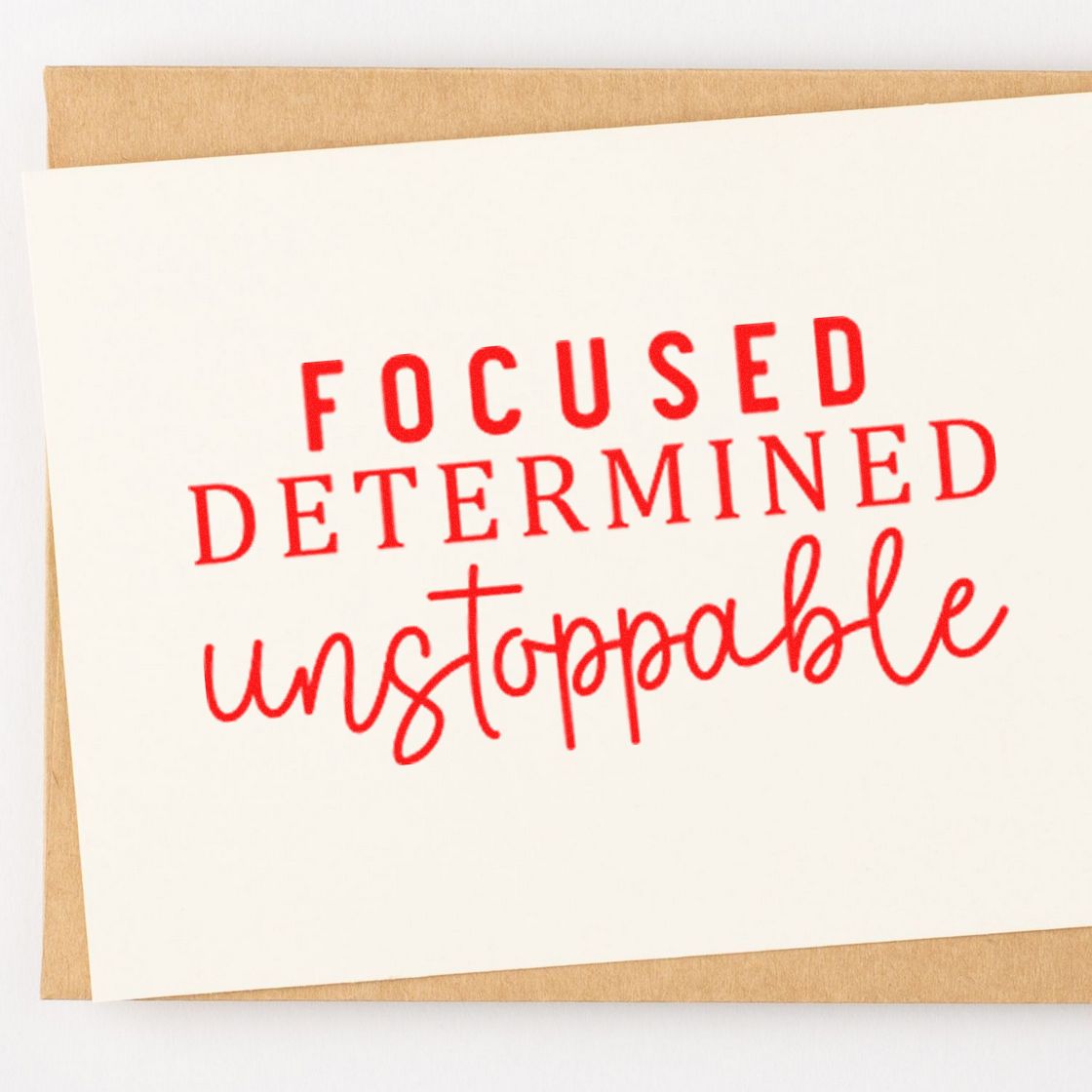 Red rubber stamp seal on white card over tan kraft paper, reading FOCUSED DETERMINED unstoppable.