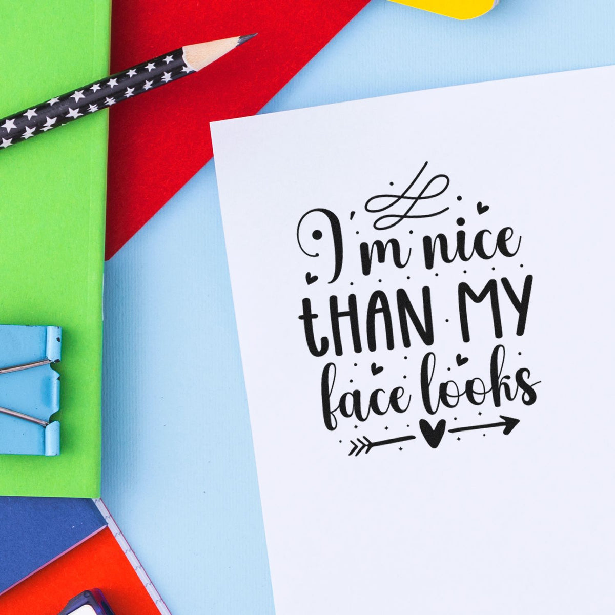 Decorative black rubber stamp on white card reading Im nicer than my face looks, with colorful paper, pencil, and clips.