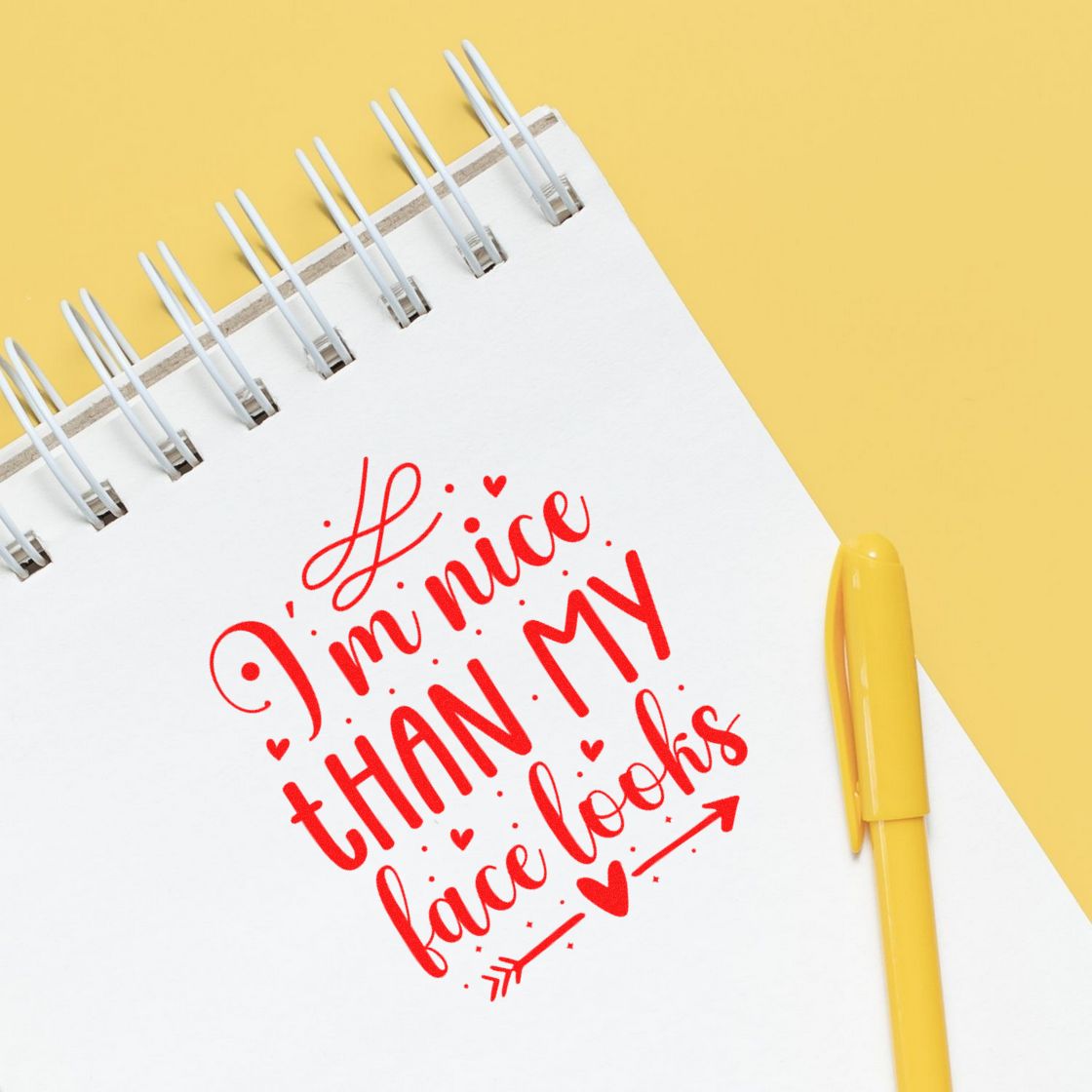 Red decorative stamp impression on white spiral notebook, with yellow pen and background, reading Im nicer than my face looks