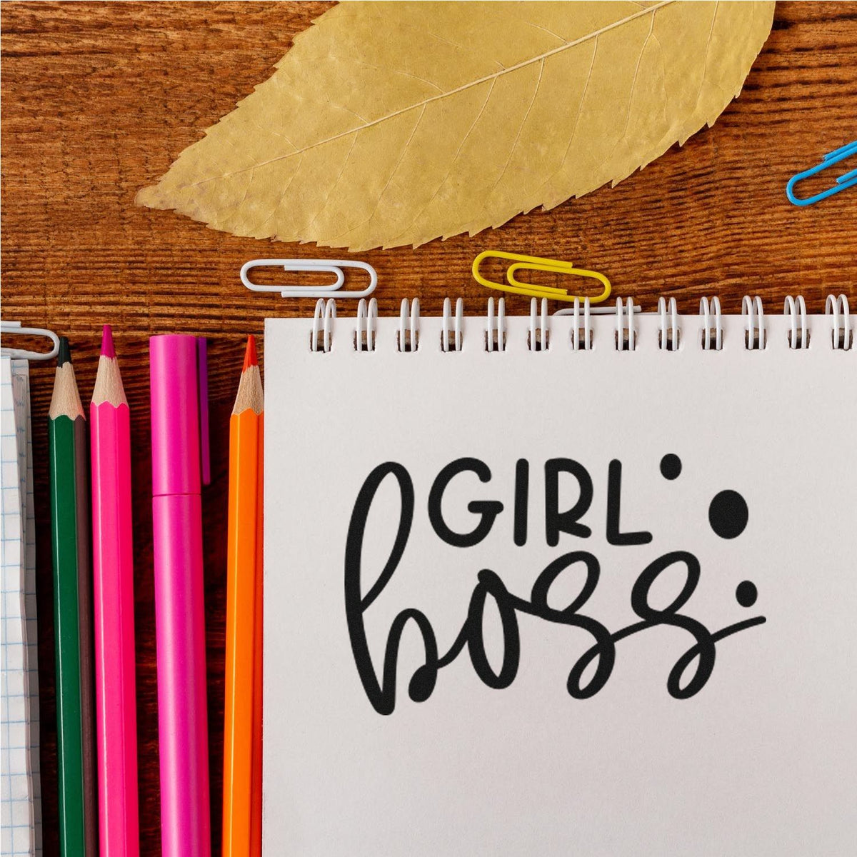 Girl Boss stamp and seal on white spiral notebook, with pink, green, orange pencils and colorful paper clips on wood.
