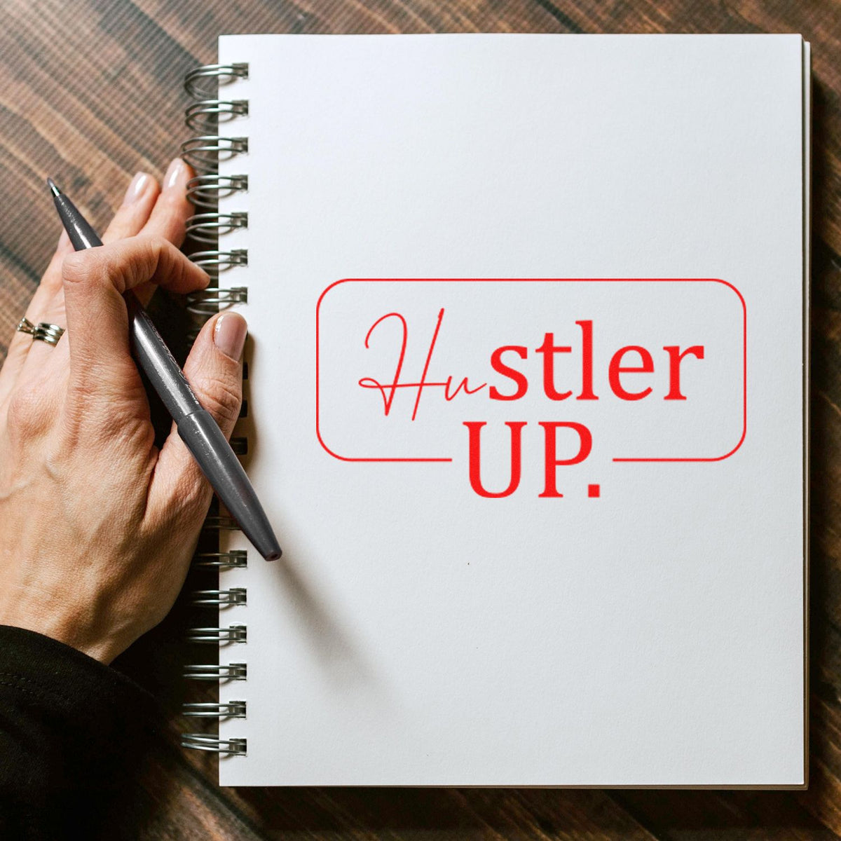 Red Hustler UP stamp and seal on white spiral notebook, held by hand with black pen.