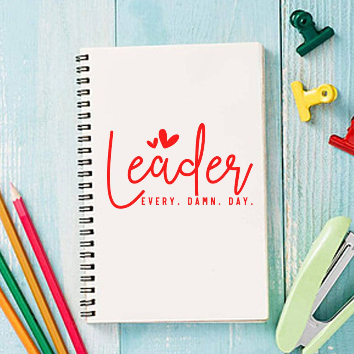 White spiral notebook with red Leader stamp and seal design, surrounded by colorful pencils and binder clips on blue wood.