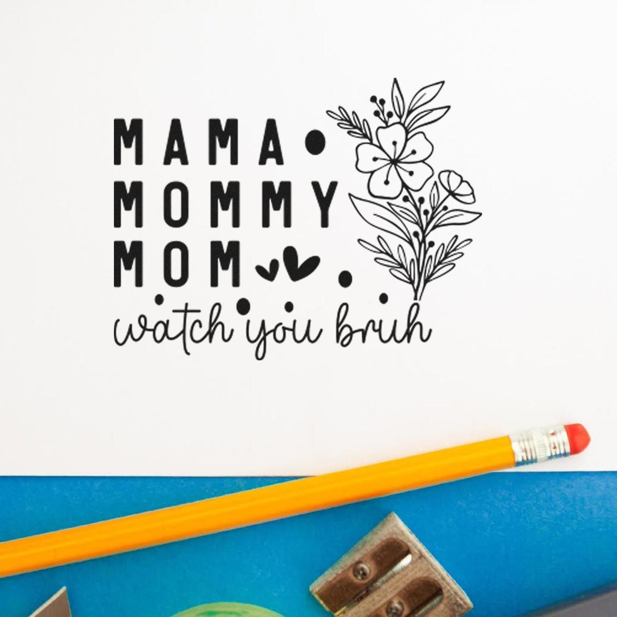 Black floral stamp seal reading MAMA MOMMY MOM watch you bruh on white paper, with a yellow pencil on blue background.