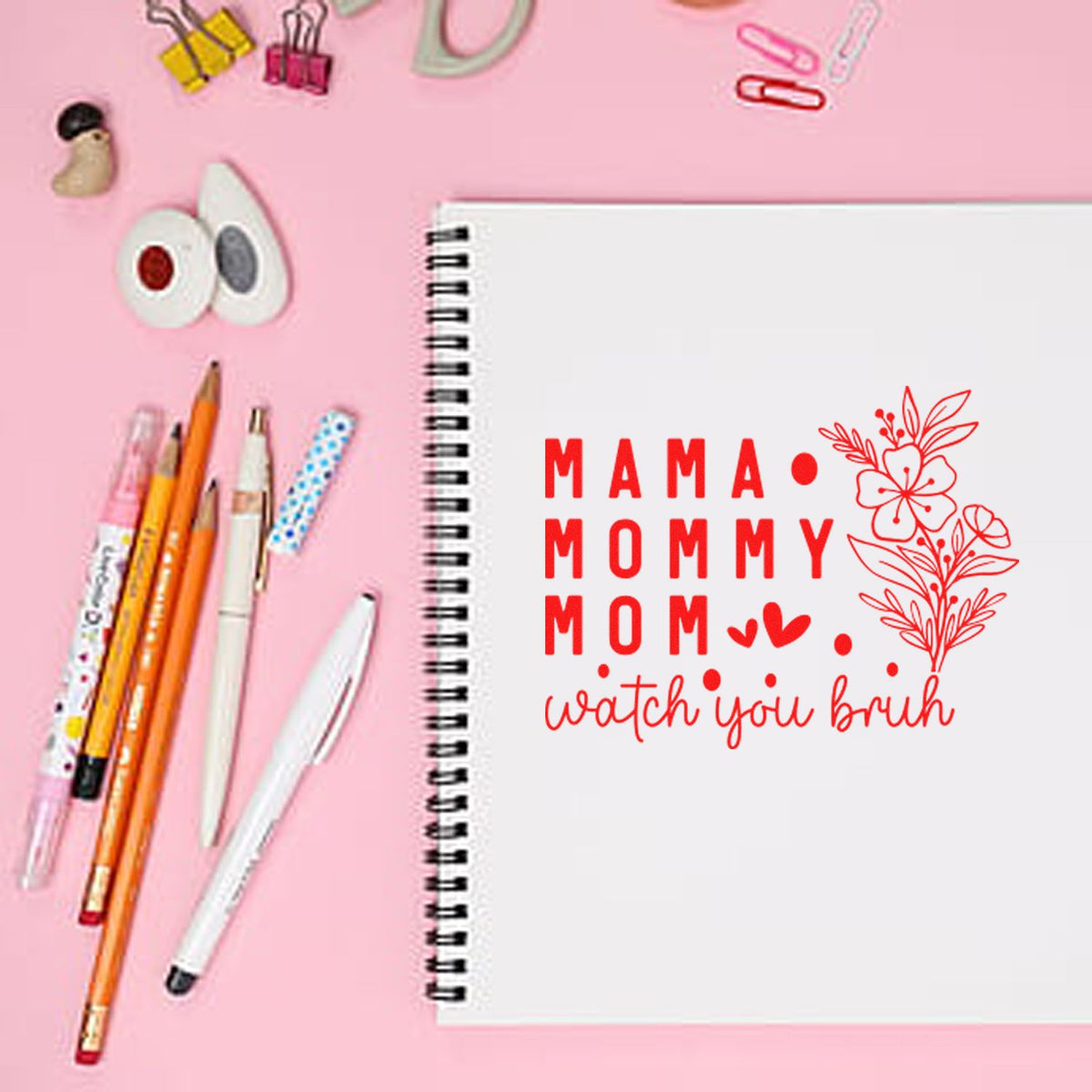 Pink notebook with a red stamp-style floral seal reading Mama Mommy Mom, watch you brush, beside pens.