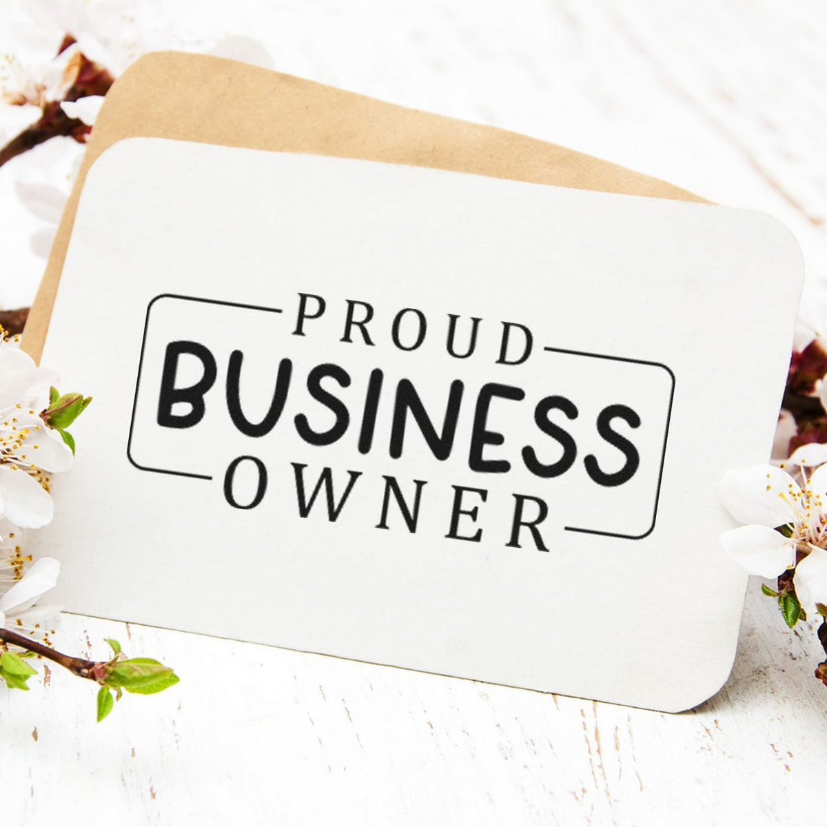 Custom business stamp and seal card with black Proud Business Owner lettering on white, over a tan envelope and white blossoms.