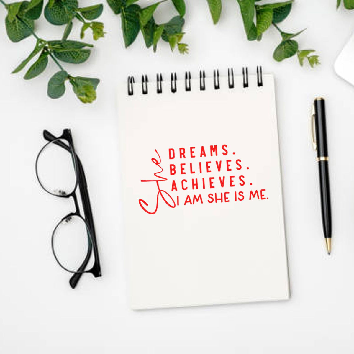 White notebook with red stamp-style seal quote, black pen, glasses, and green leaves on a clean desk.
