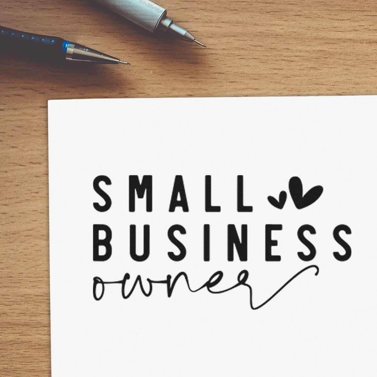 Black stamp and seal reading Small Business owner on white paper, with pens on a wood desk.