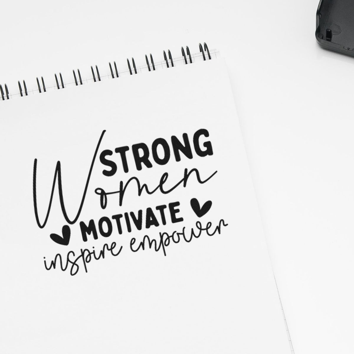 Black motivational stamp/seal design on a white spiral notebook, reading Strong Women Motivate Inspire Empower with hearts.