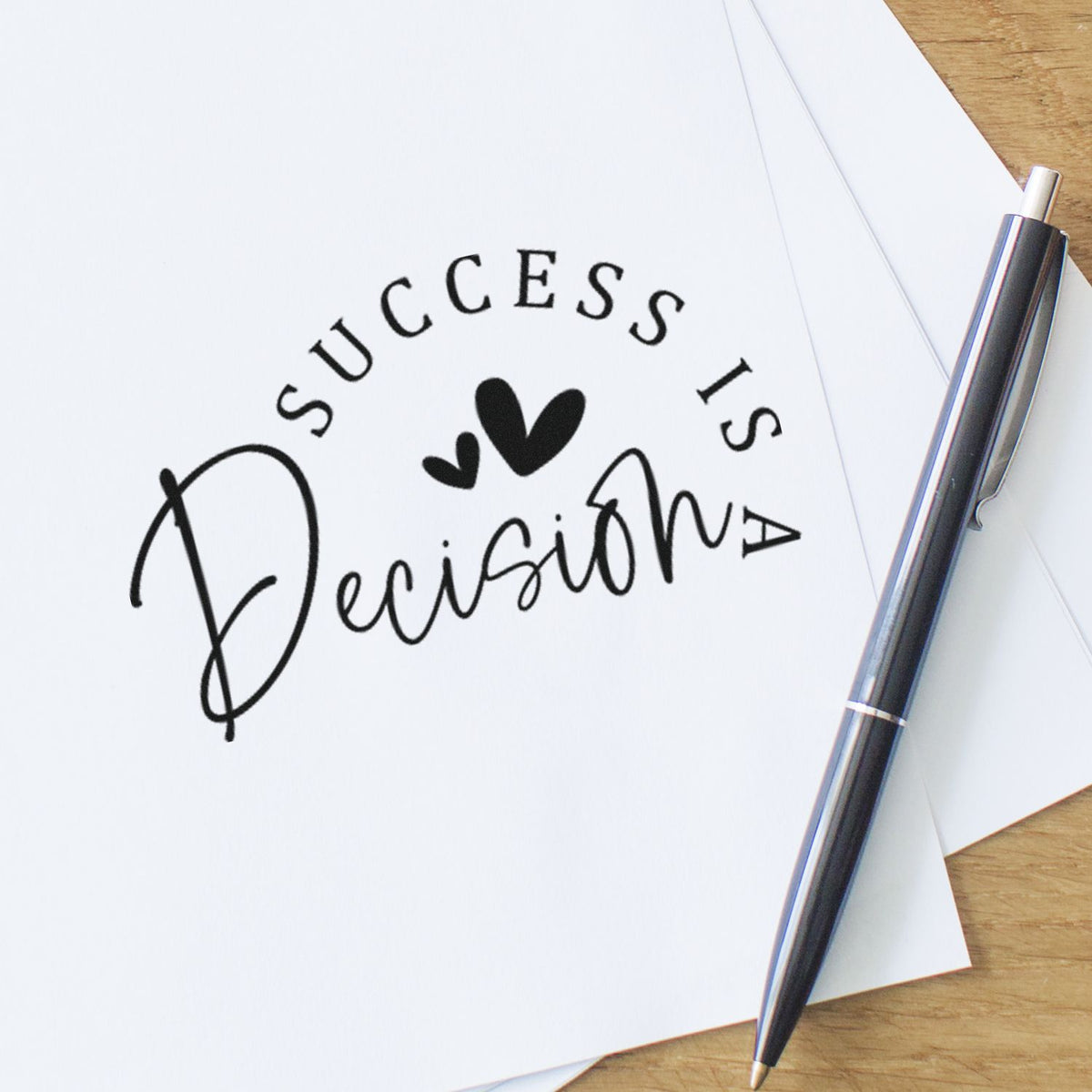 Black Success Is A Decision stamp seal with hearts on white paper, beside a silver-and-black pen on a wood desk.