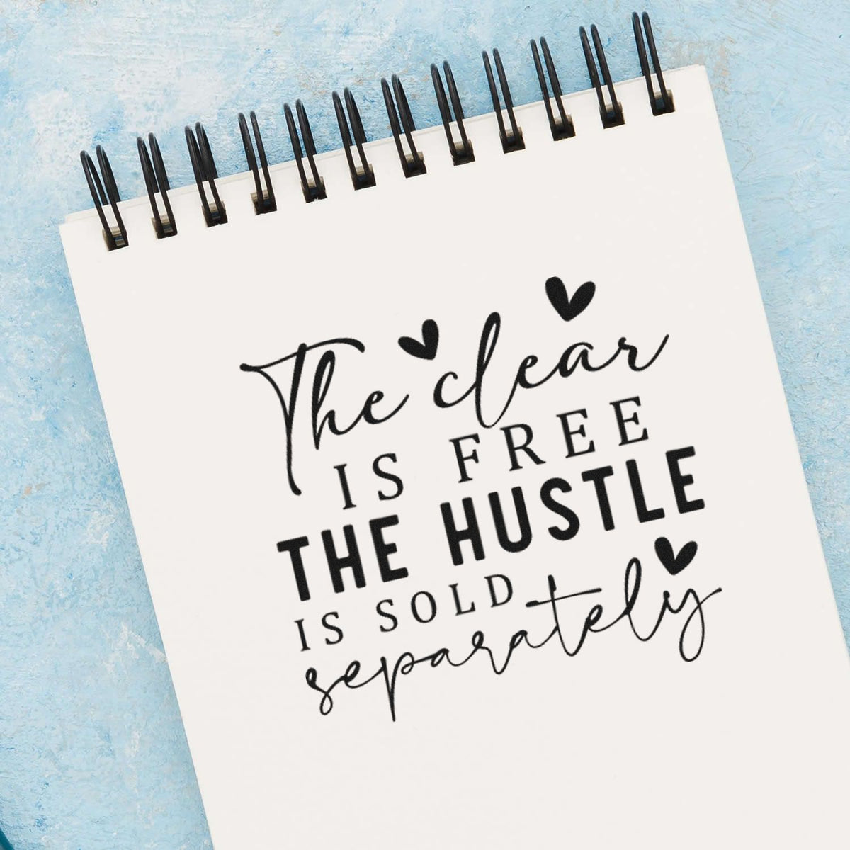 White spiral notebook featuring a black stamp/seal quote design, hearts, and bold lettering on a light blue background.