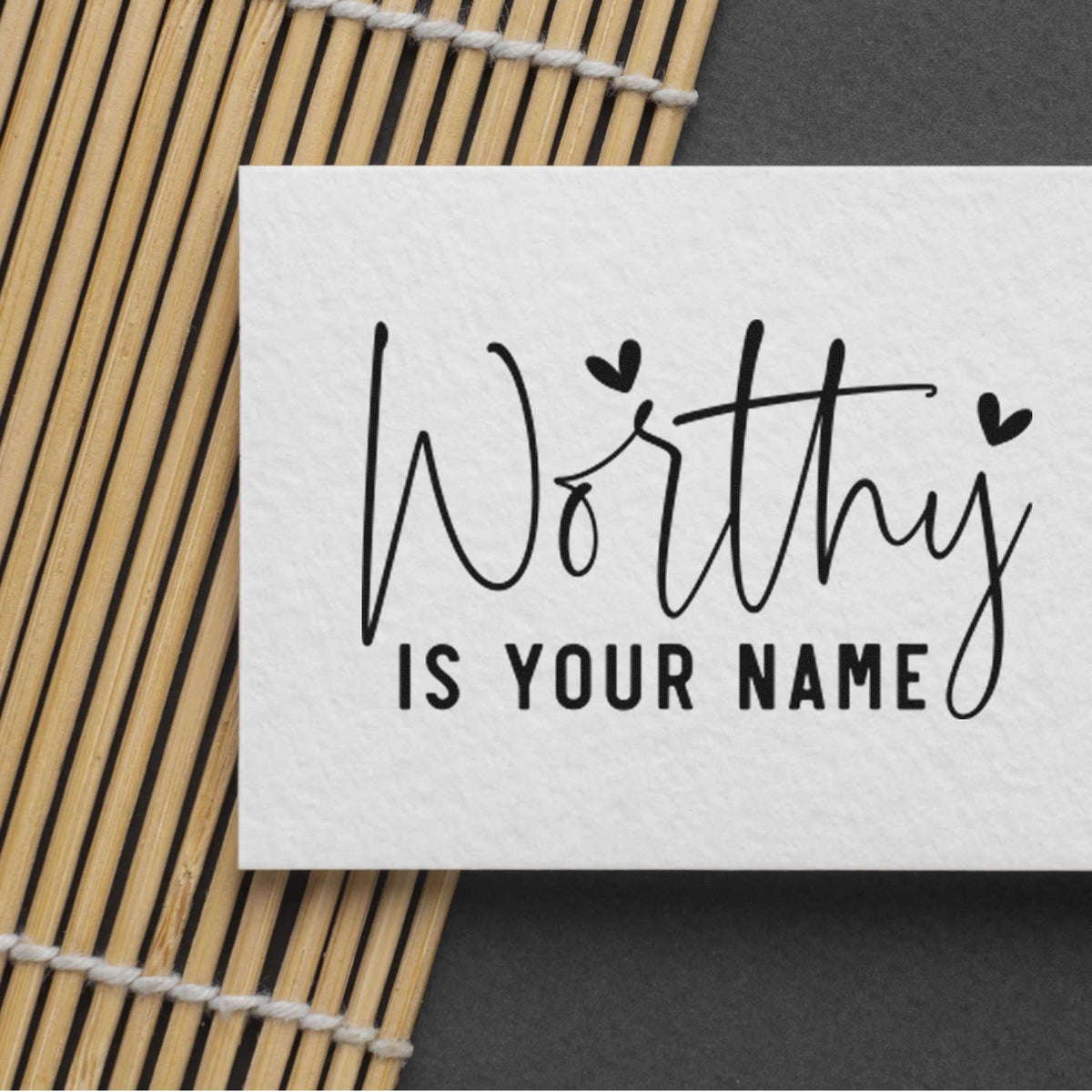 Personalized black stamp and seal reading Worthy is your name on a white card over a bamboo mat and dark background