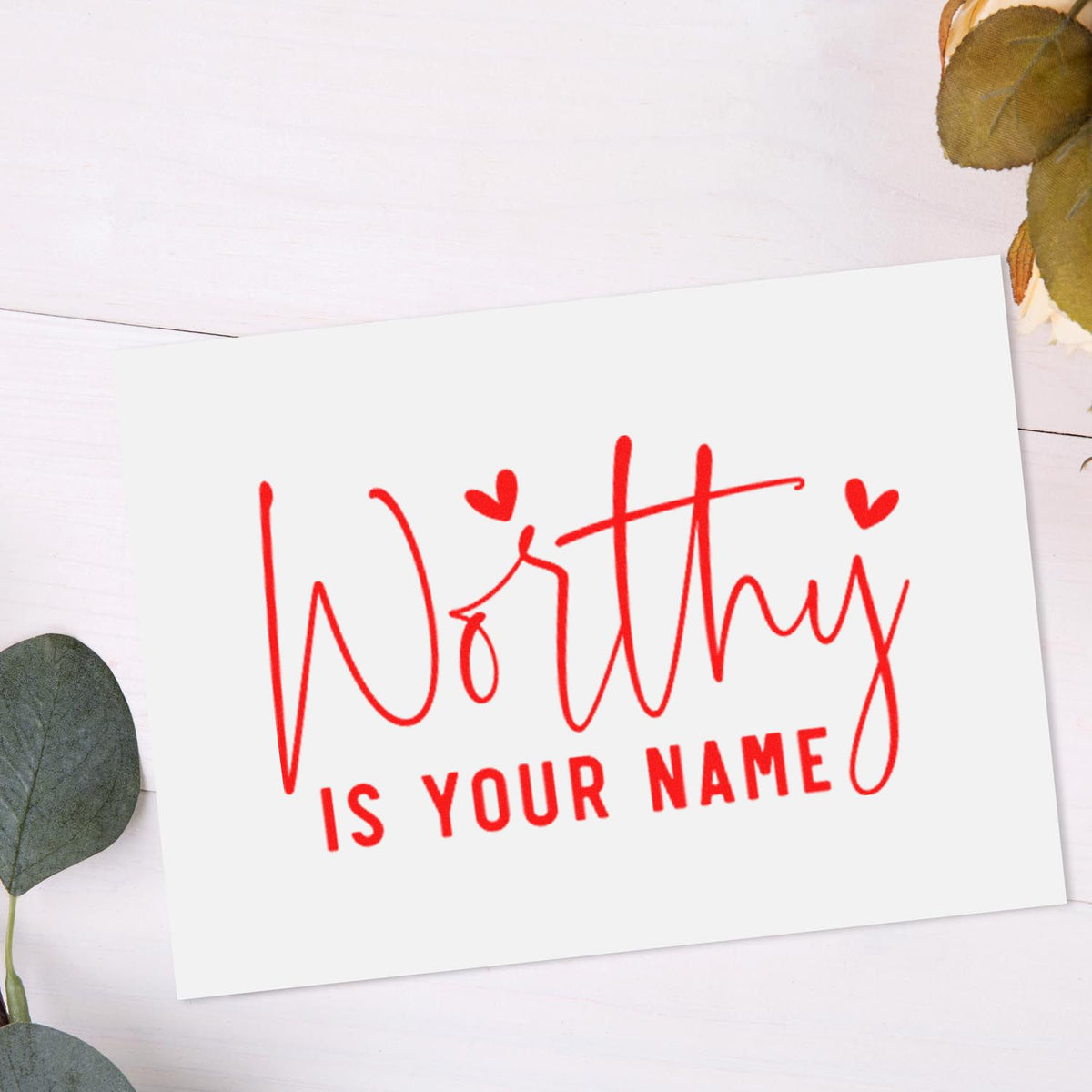 Red custom stamp and seal reading Worthy is your name on a white card with heart accents and eucalyptus leaves.