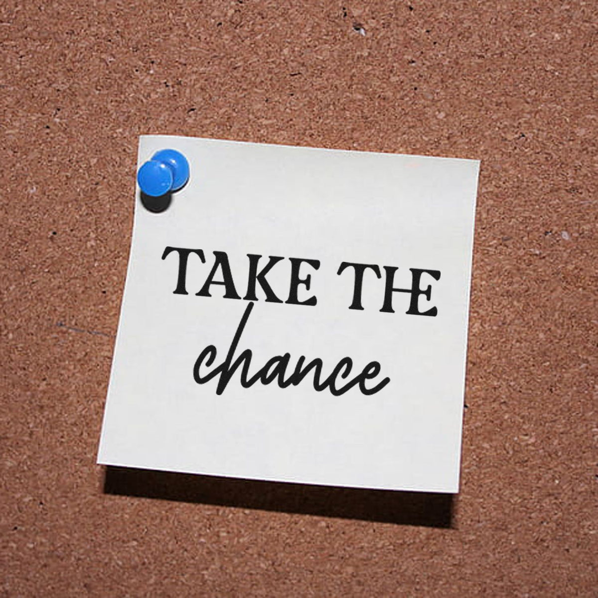 White note pinned with blue pushpin on cork board, featuring black take-the-chance stamp and seal lettering.
