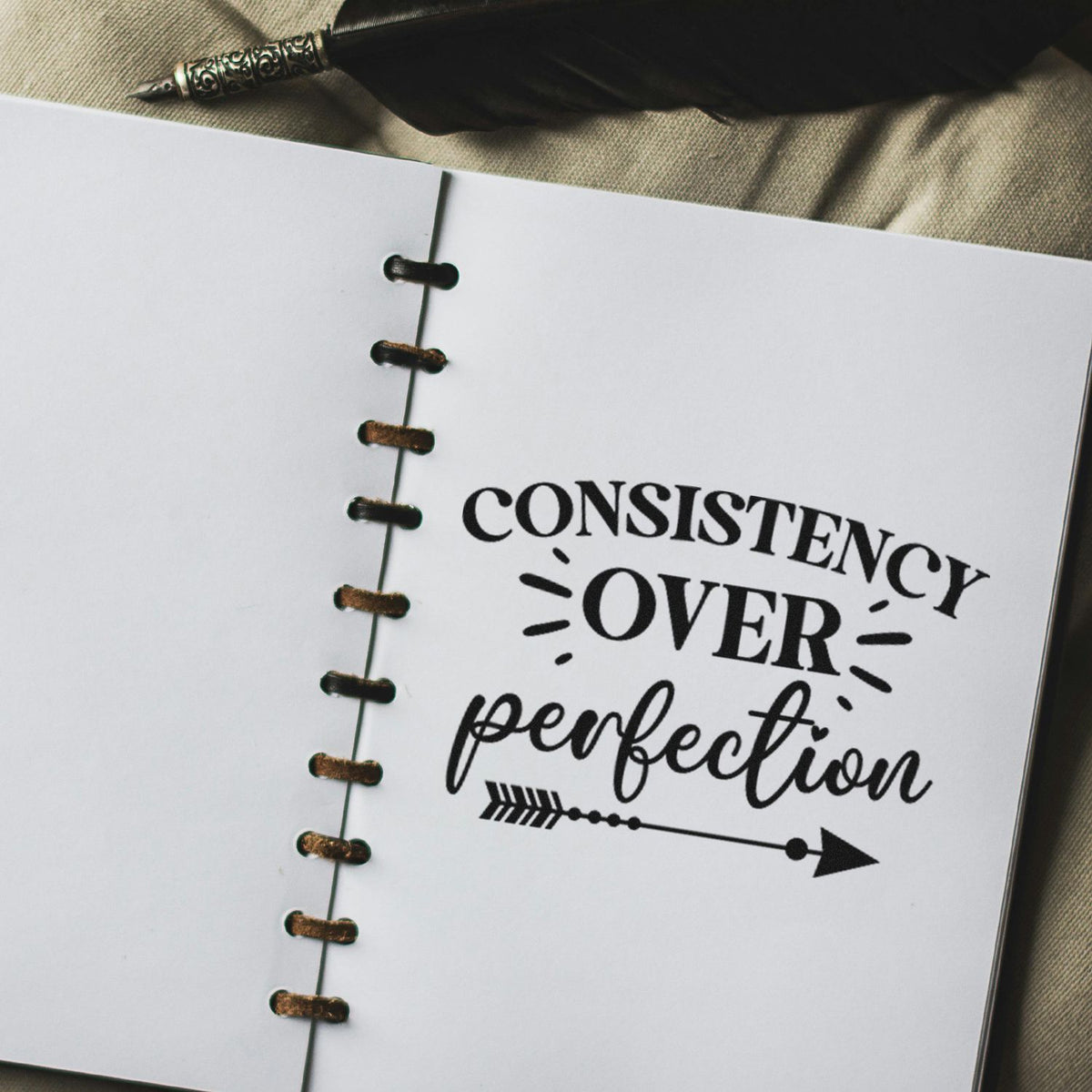 Black stamp and seal design on white notebook page reading Consistency over perfection with arrow and binder holes.