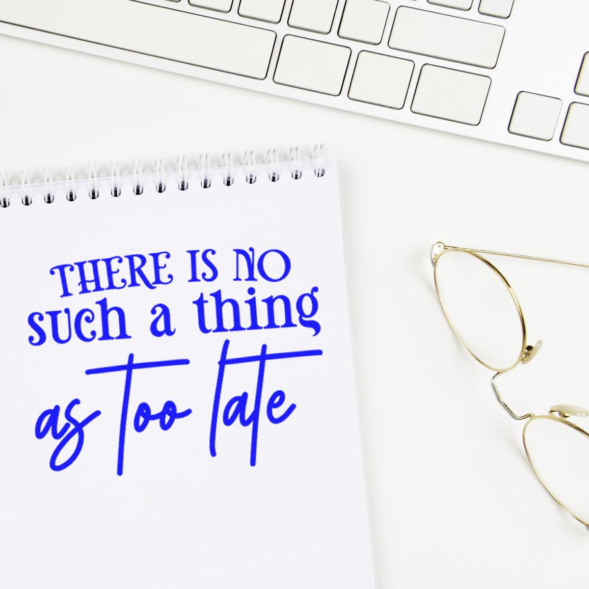 Blue stamp seal on white notebook reads There is no such thing as too late, beside a white keyboard and gold glasses.